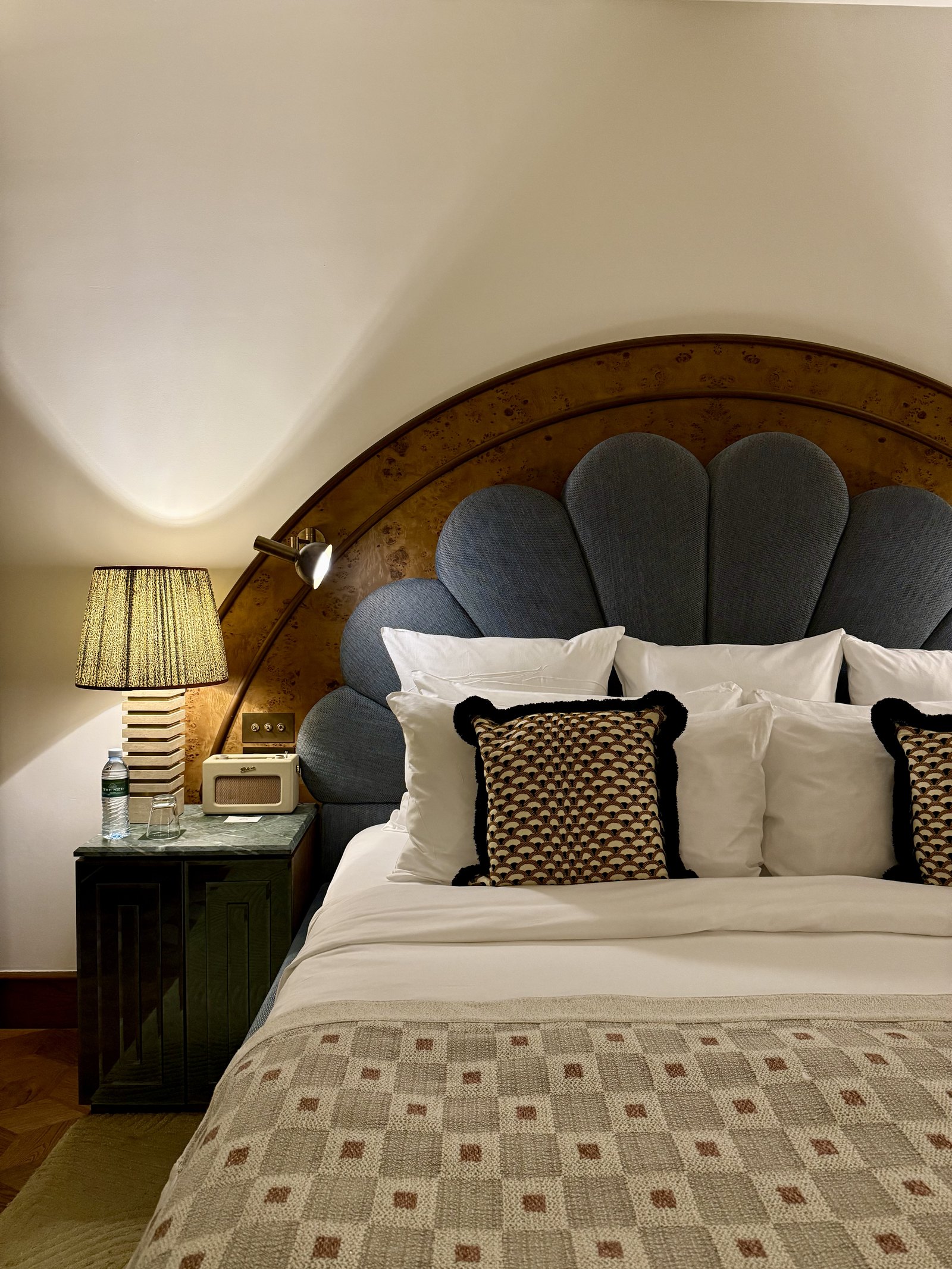 A bedroom at The Ned Doha with a grey velvet shell headboard, patterned cushions and a woven bedcover, a shaded lamp beside it