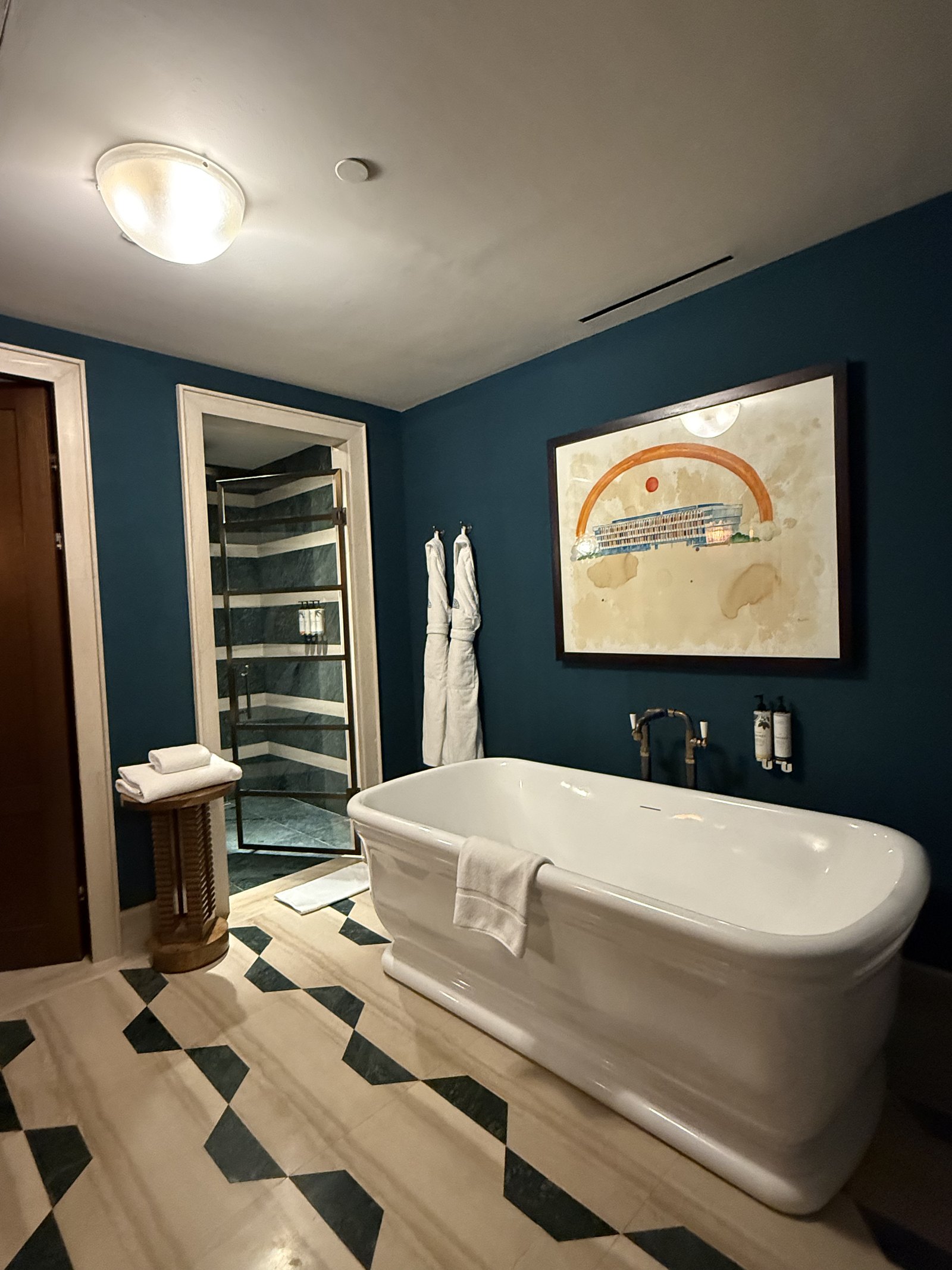 A bathroom at The Ned Doha with deep blue walls, a freestanding white tub and a graphic black and cream marble floor
