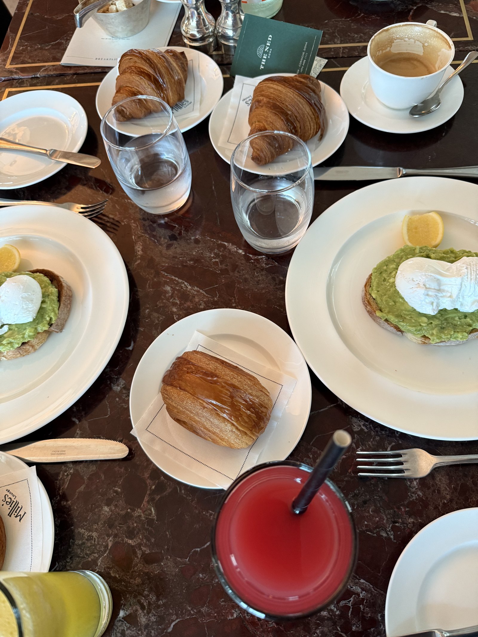 A breakfast table from above at The Ned Doha with croissants, avocado toast with a poached egg, coffee and a glass of watermelon juice on dark marble