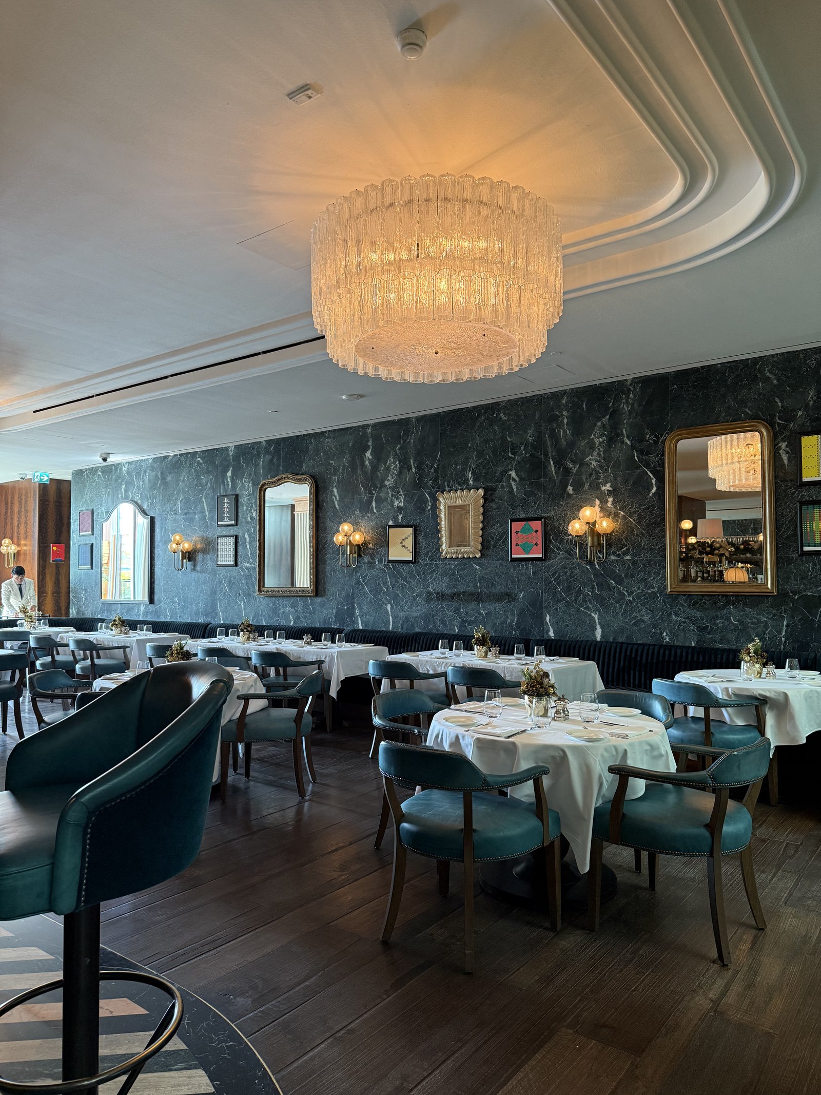 The dining room at Cecconi's in The Ned Doha, dark green marble walls hung with pictures, teal chairs and white linen under a glass chandelier