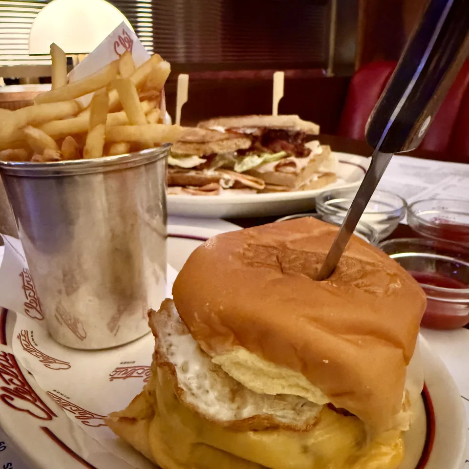 A burger with a fried egg on a plate beside a metal cup of fries, on a red leather banquette table