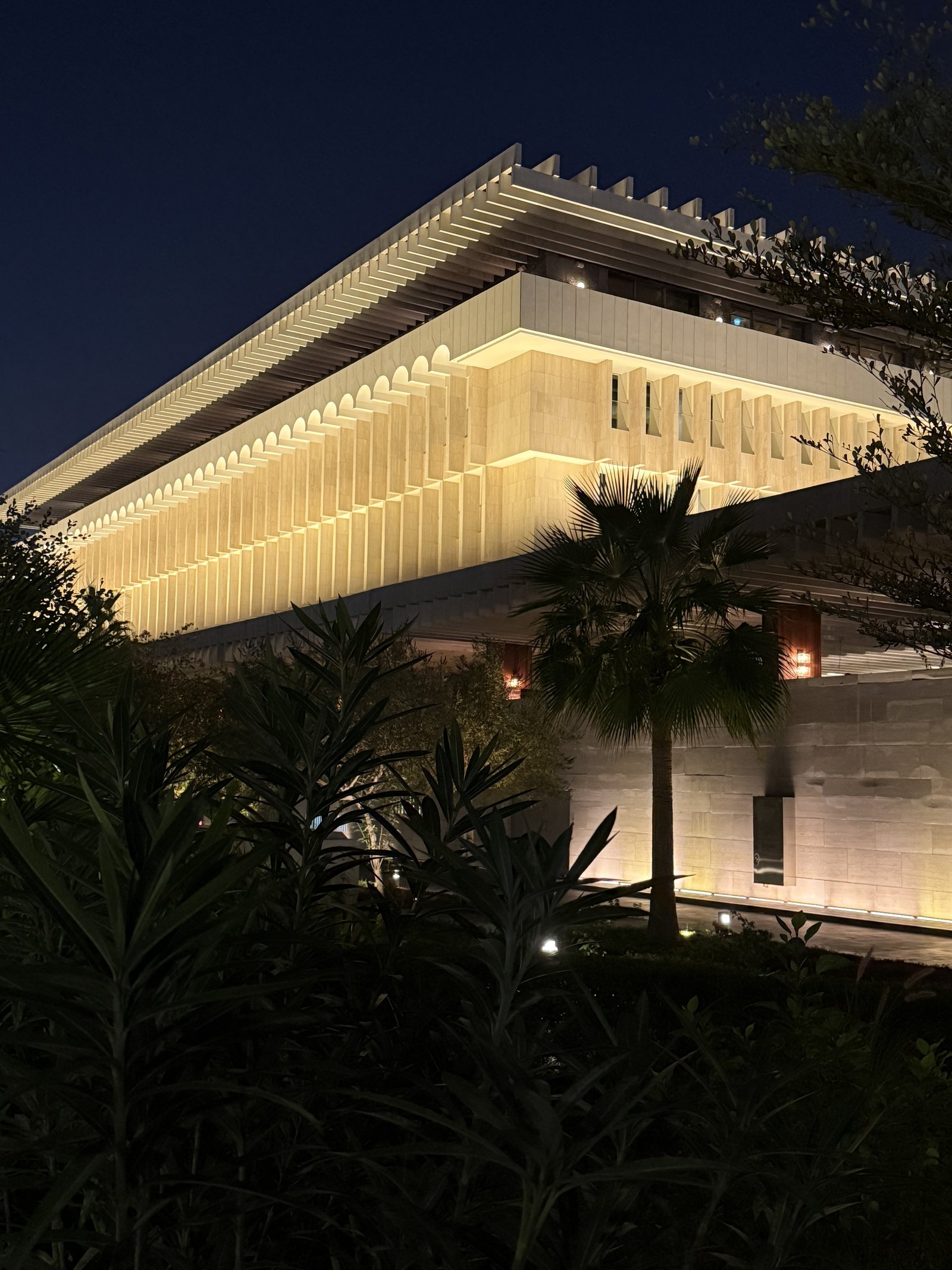 The former Ministry of Interior in Doha at dusk, a long concrete cantilever lit from below over a dark colonnade, with palms in front of it