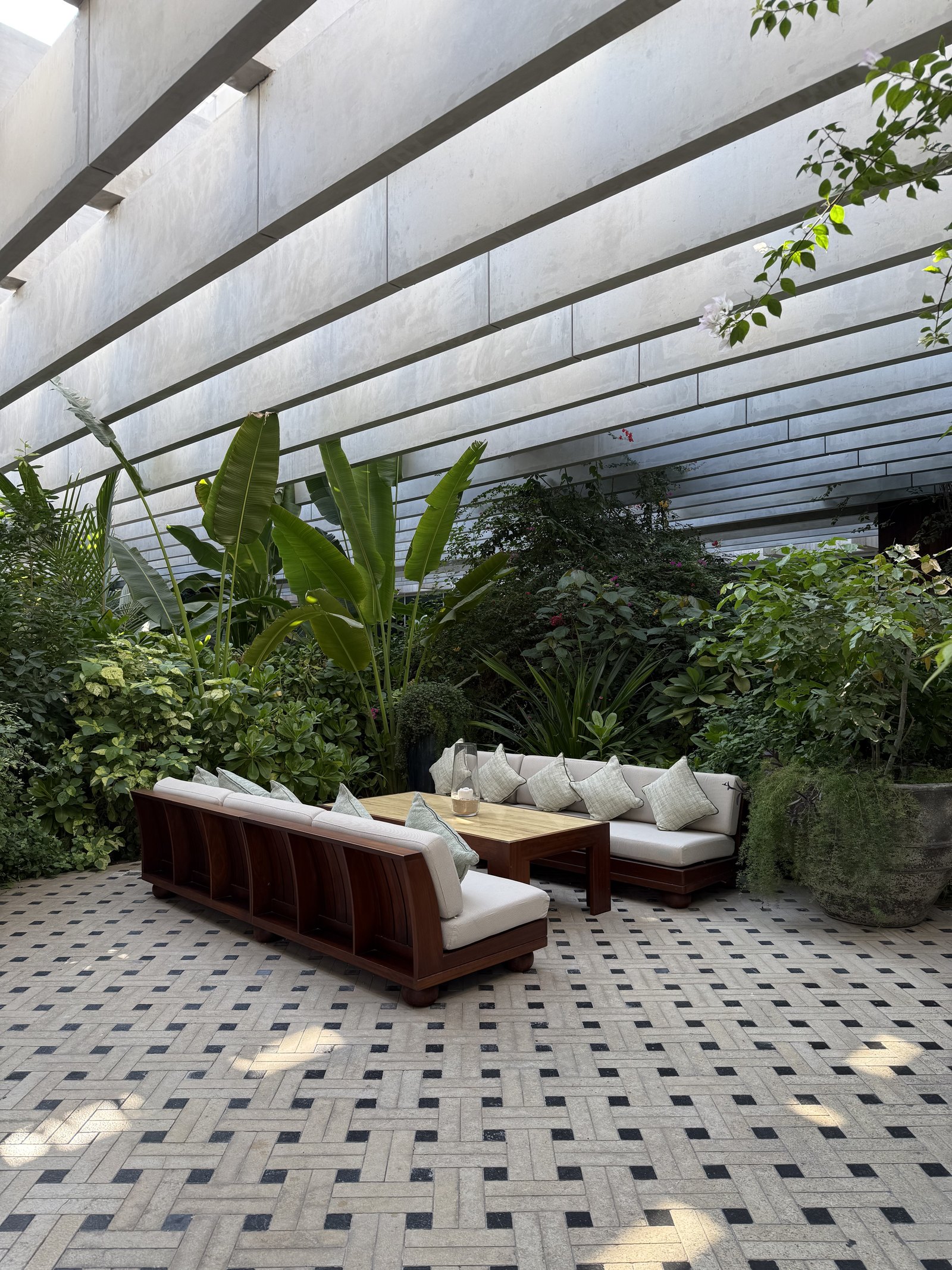 A garden seating area at The Ned Doha under deep concrete beams, with low sofas, banana plants and a patterned stone floor