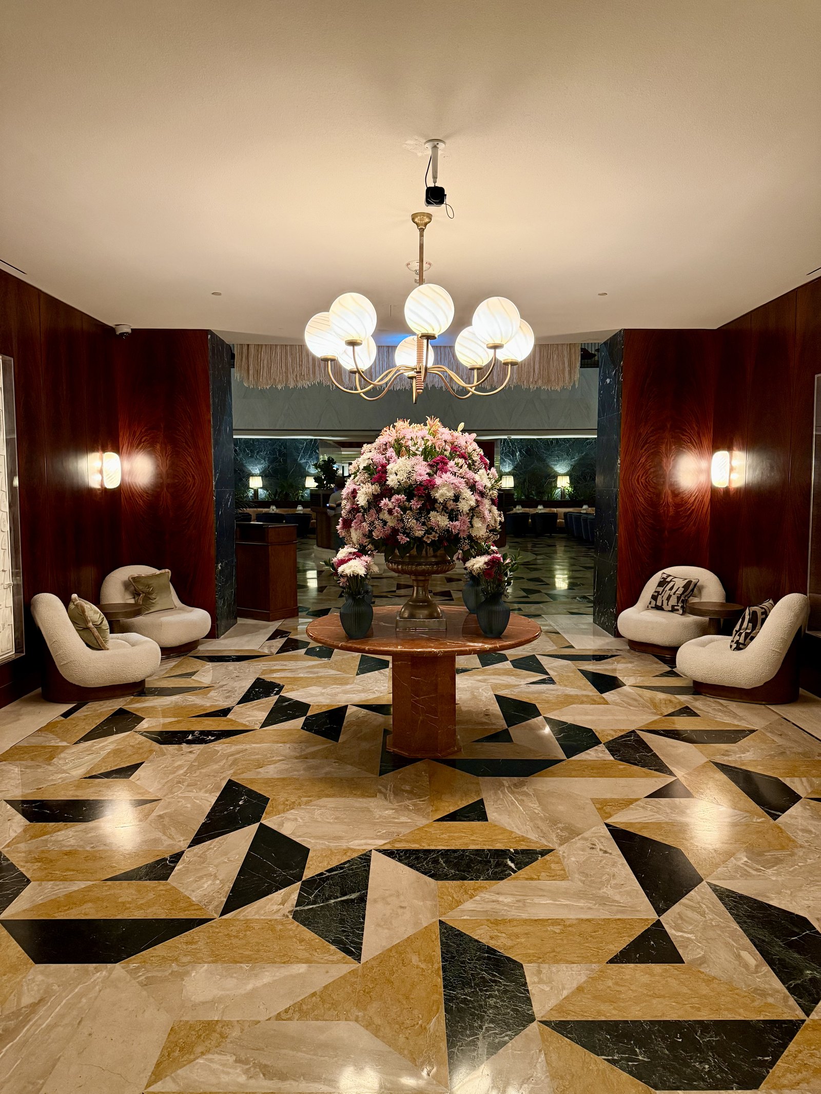 The lobby at The Ned Doha, a floor of inlaid marble in a radiating star pattern, a round table with pink flowers under a glass chandelier and curved white sofas