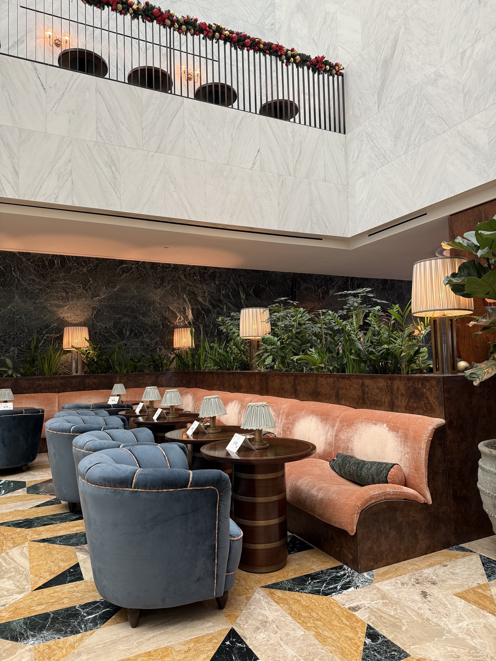 A lounge at The Ned Doha with blue velvet tub chairs, a pink banquette and potted plants on a patterned marble floor