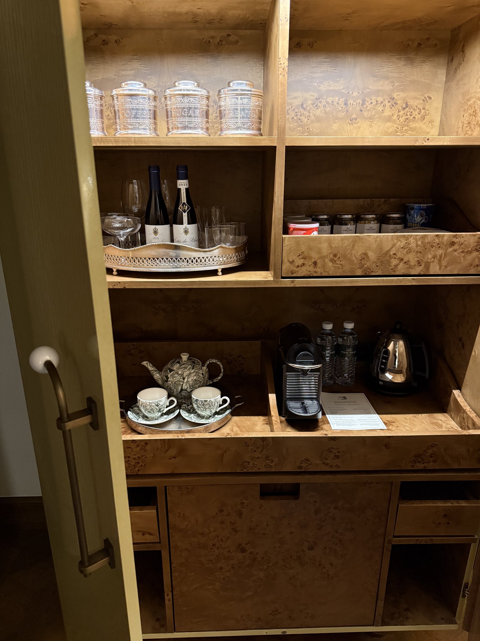 An open burl wood cabinet in a room at The Ned Doha holding glasses, a coffee machine, a teapot and two cups