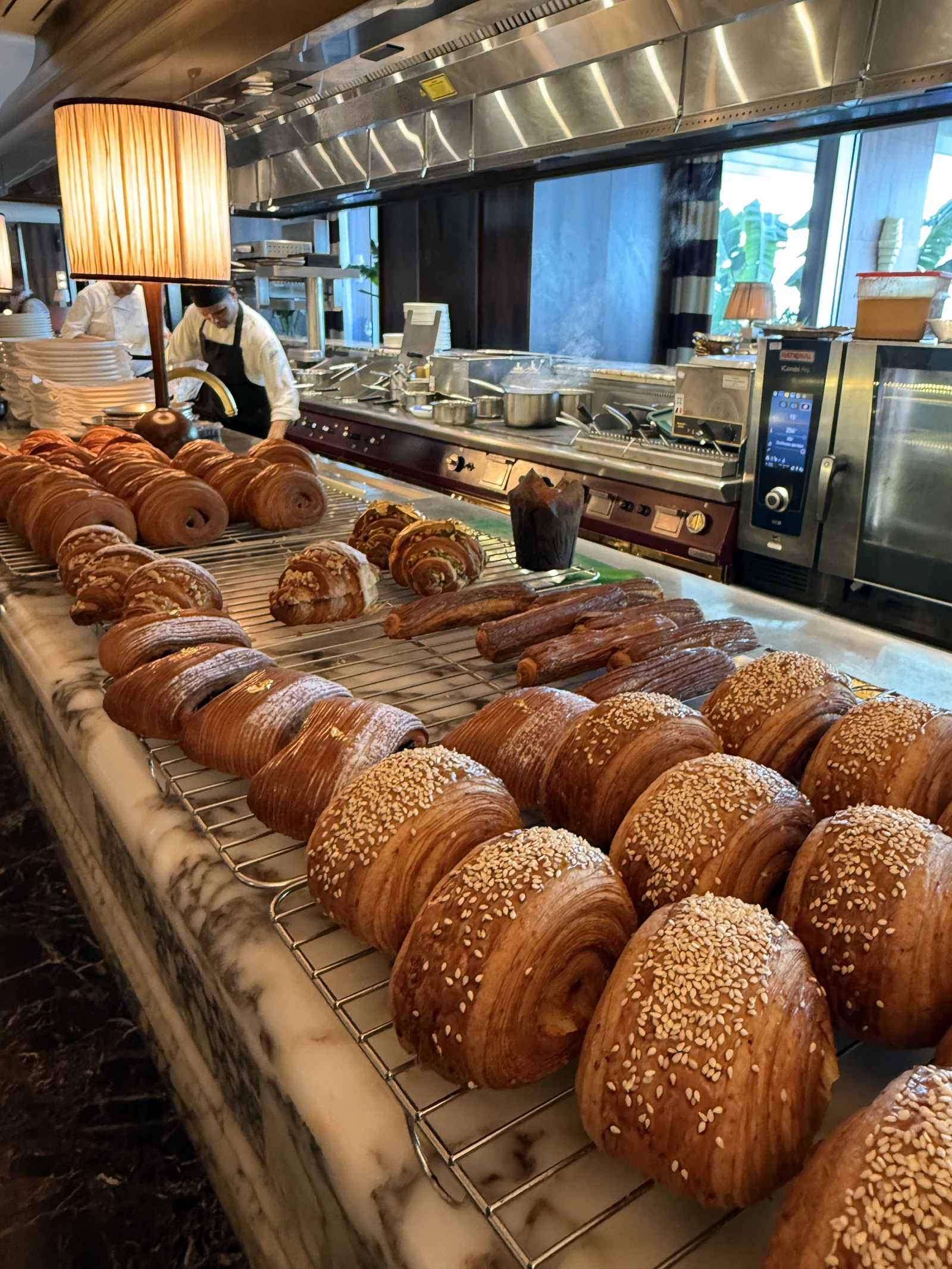 A marble counter at The Ned Doha laid with rows of croissants and sesame rolls on wire racks, a cook working at the pass behind