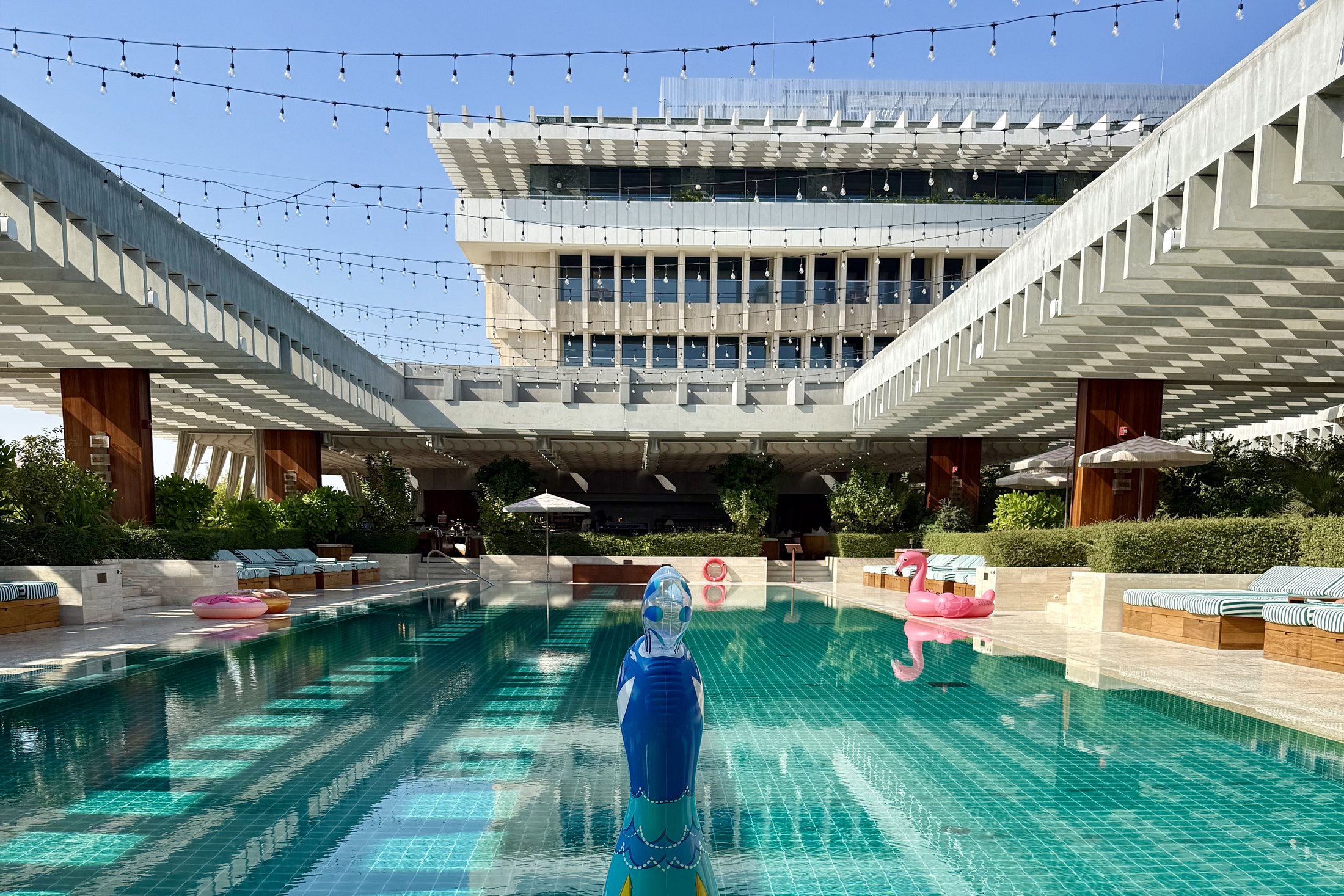 The thirty metre pool at The Ned Doha in daylight with the full concrete facade of the former ministry standing over the far end, striped loungers down both sides and festoon lights strung overhead