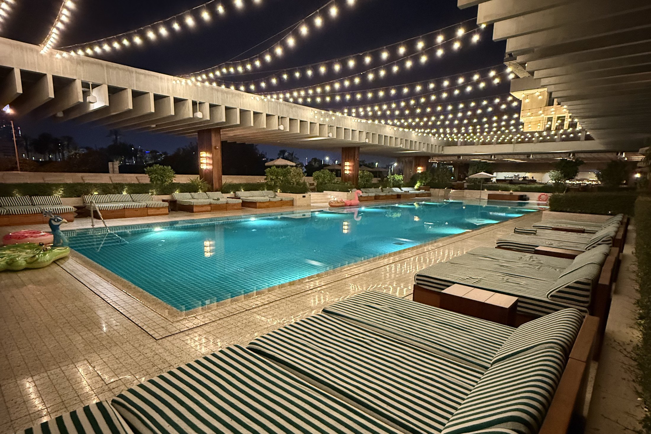 The pool at The Ned Doha at night, lit turquoise under strings of festoon lights, with a long run of green and white striped daybeds along the near side and the concrete colonnade above
