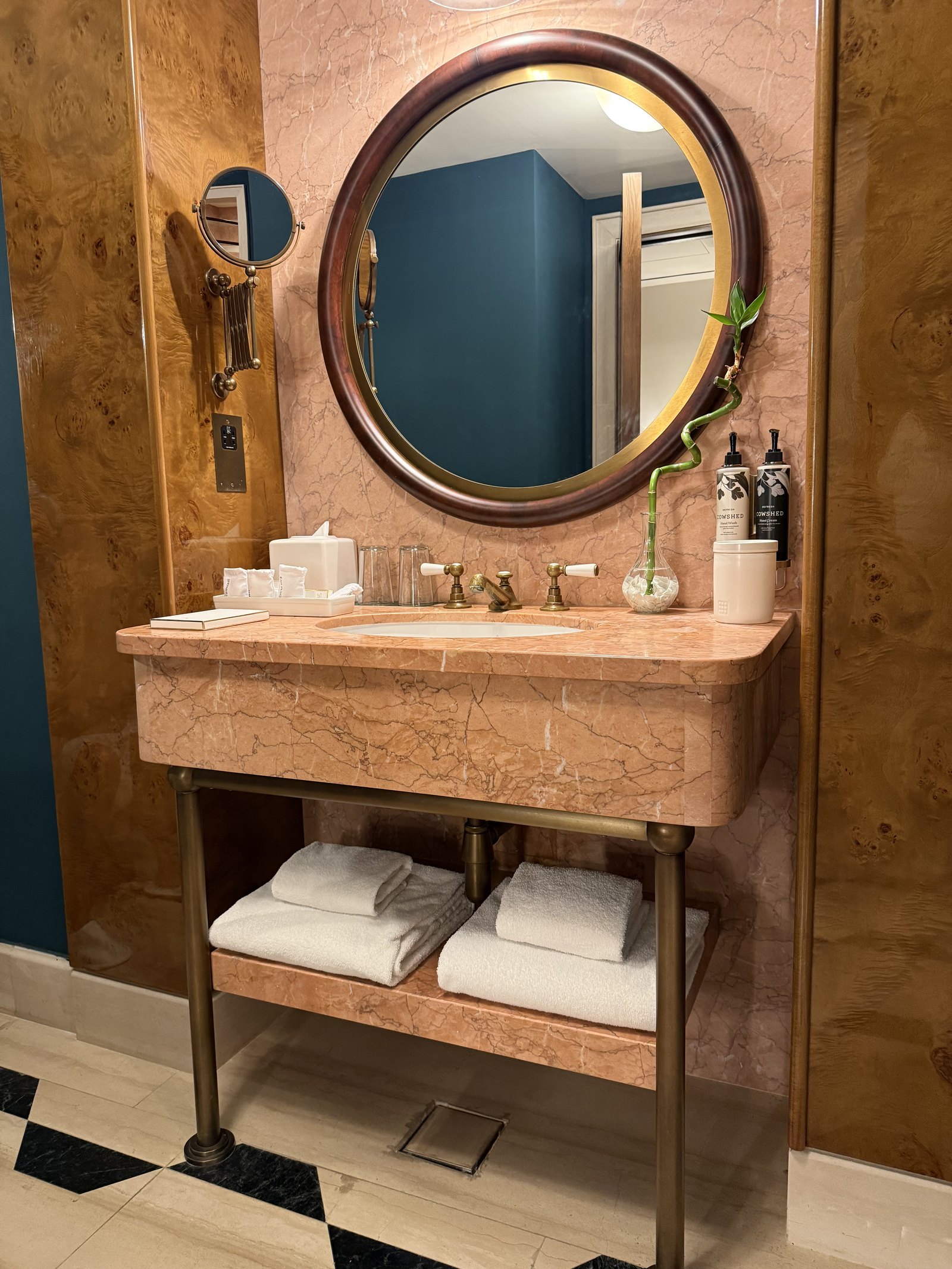 A bathroom vanity at The Ned Doha in rose marble with an oval brass mirror above it and a deep blue wall behind