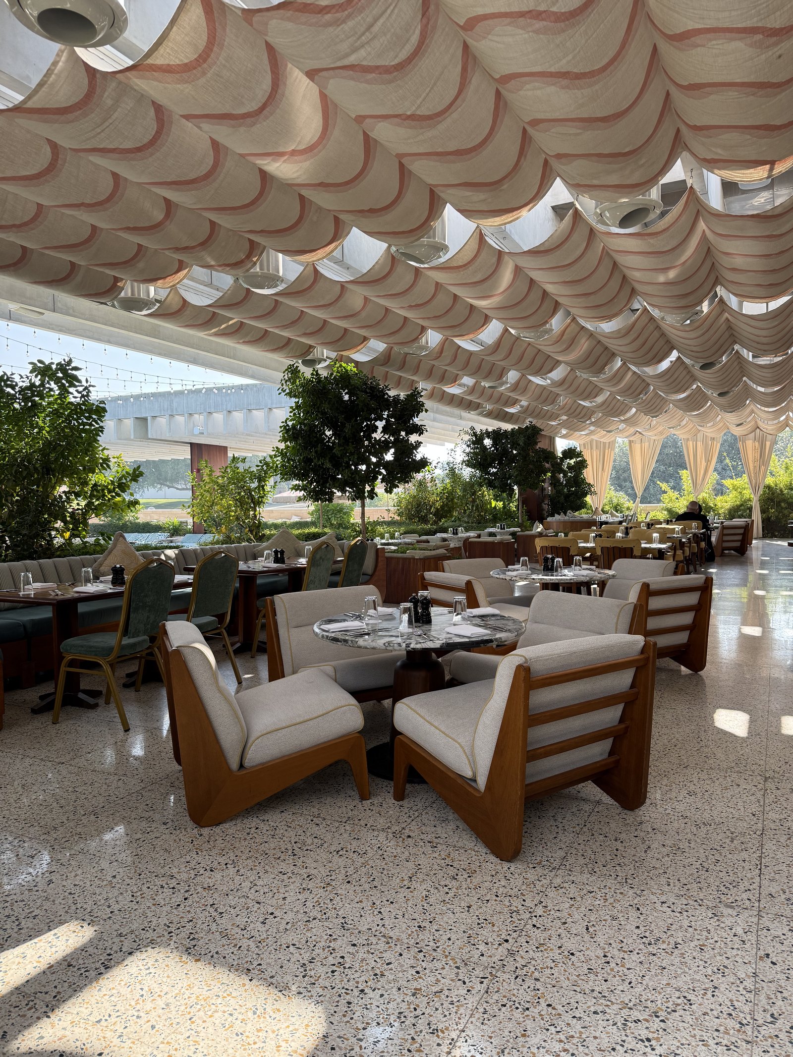 An outdoor restaurant at The Ned Doha under a canopy of overlapping pale scallops, with slatted timber armchairs, potted trees and a terrazzo floor