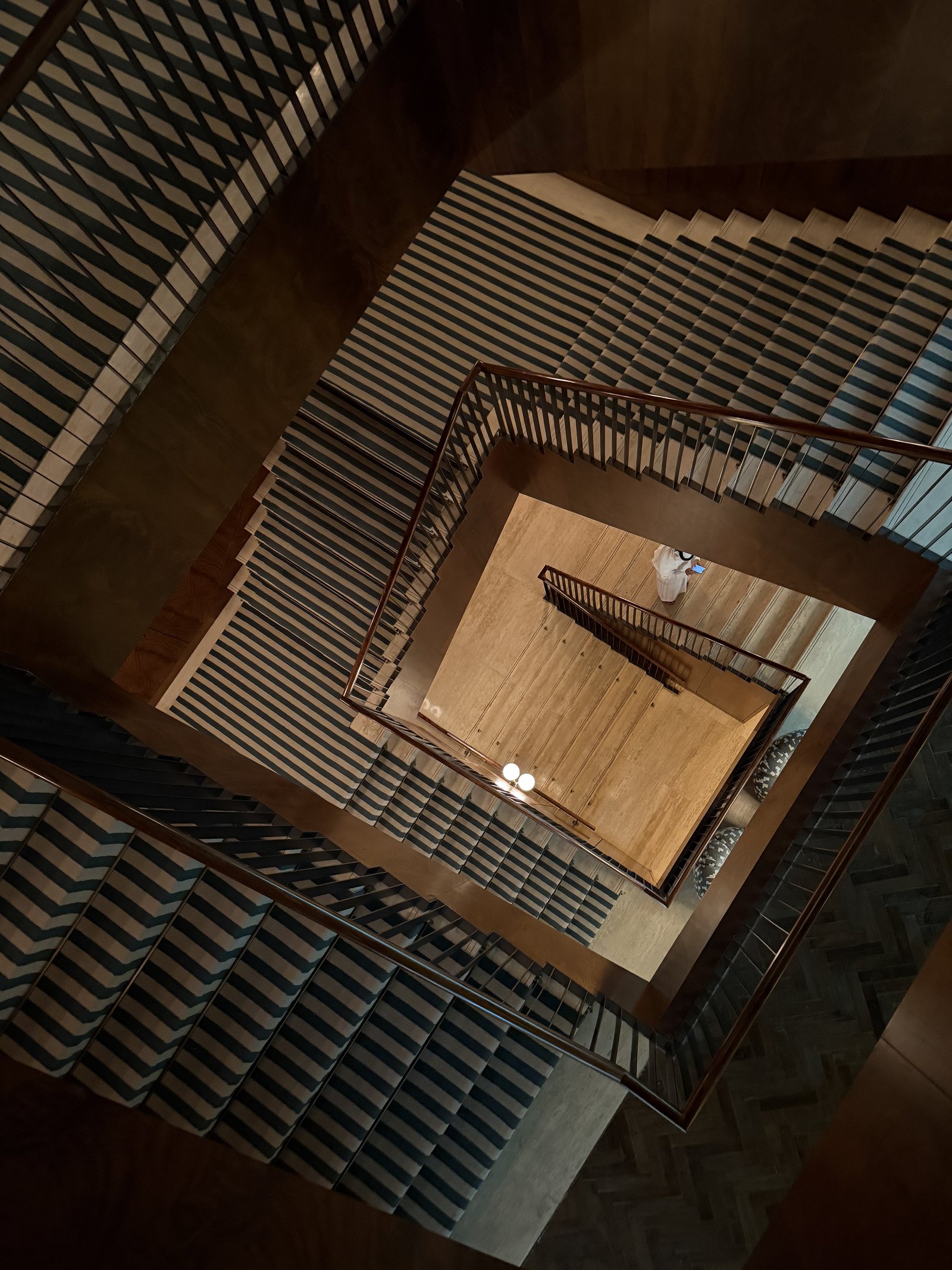 A stairwell at The Ned Doha from directly above, the square flights running down around a lit well with striped carpet
