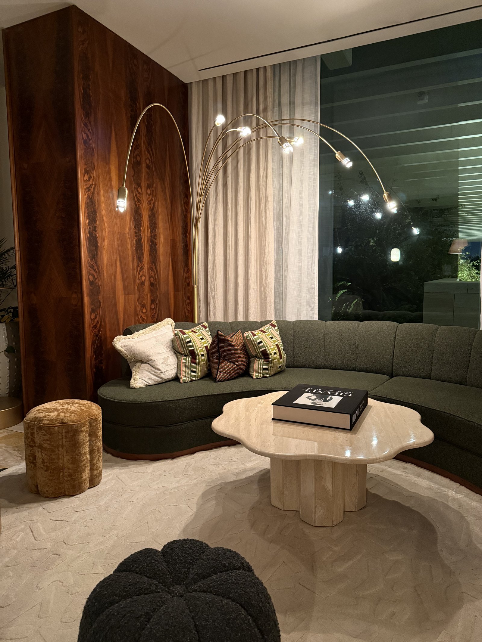 The sitting end of a suite at The Ned Doha with a curved green sofa, a scalloped travertine table, an arc lamp and walnut panelling, the glass running the full width