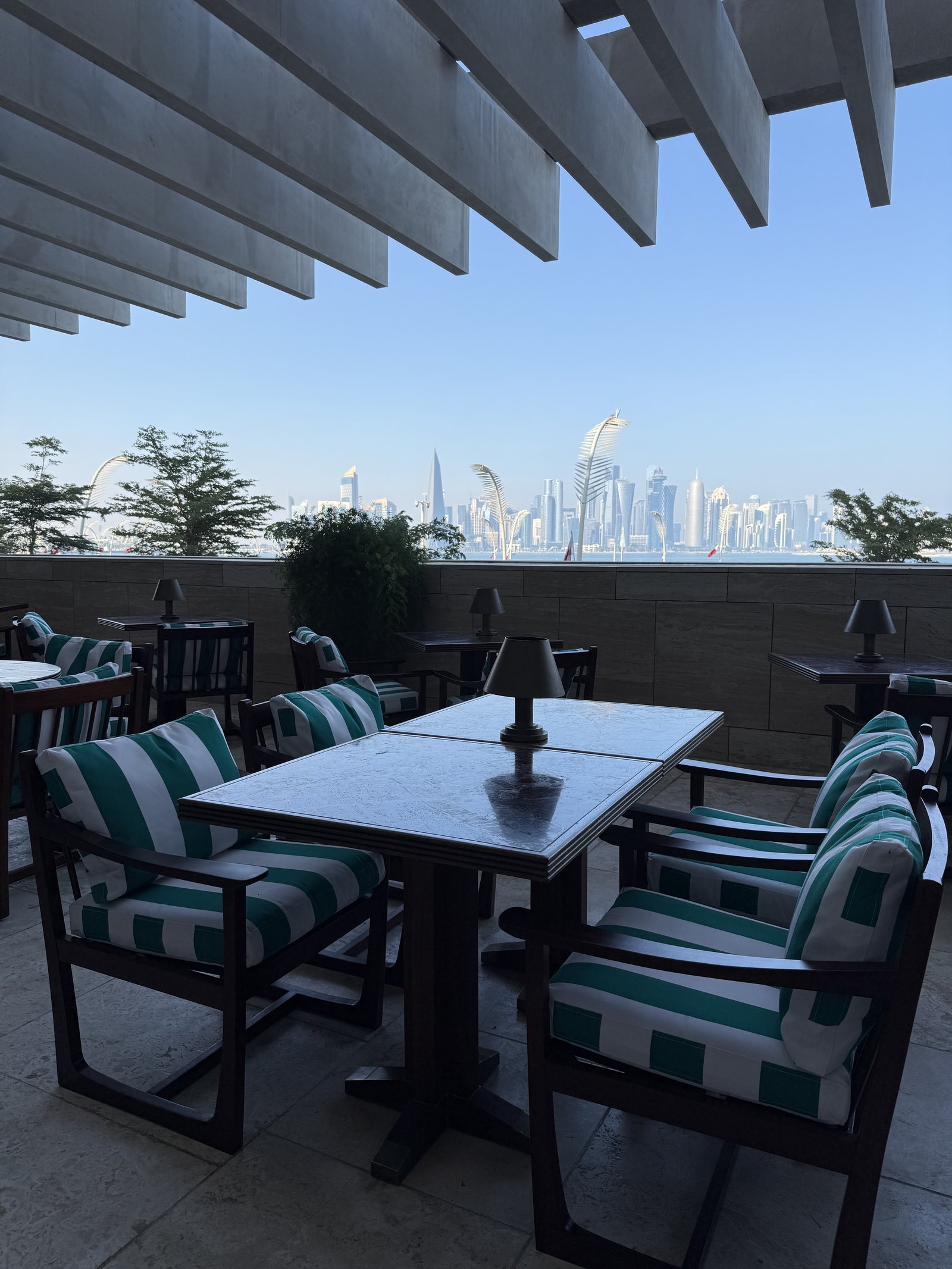 A terrace at The Ned Doha with green and white striped chairs around a dark table, the towers of West Bay on the skyline beyond the wall