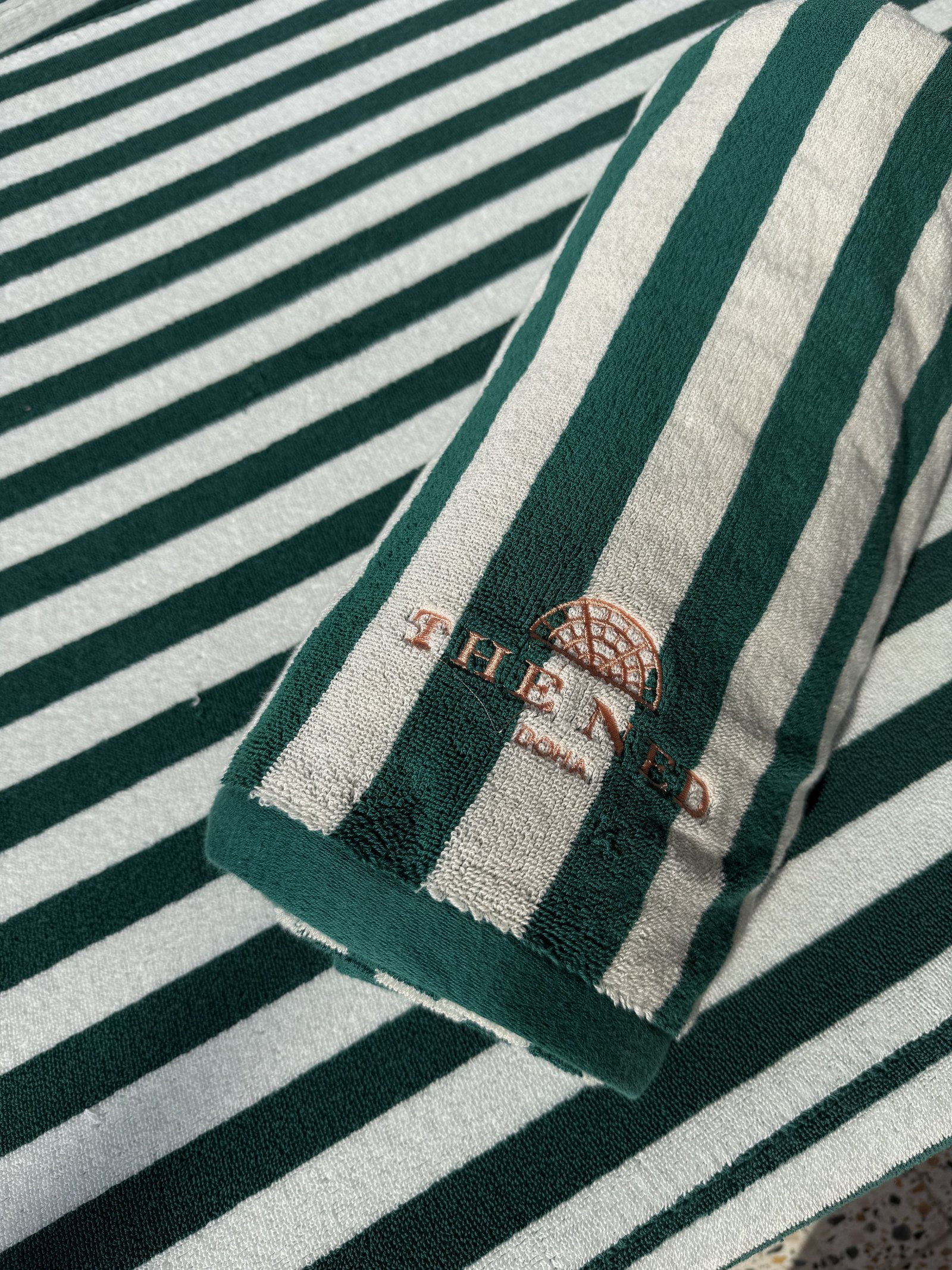 A rolled green and white striped pool towel with The Ned embroidered on it in rust thread