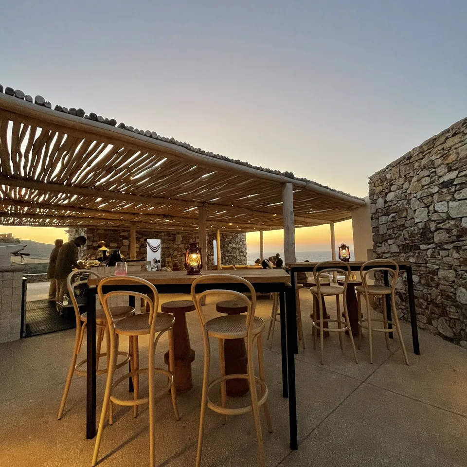 The open-air bar at The Rooster at sundown, bentwood stools along a counter under a timber roof with the light going orange behind
