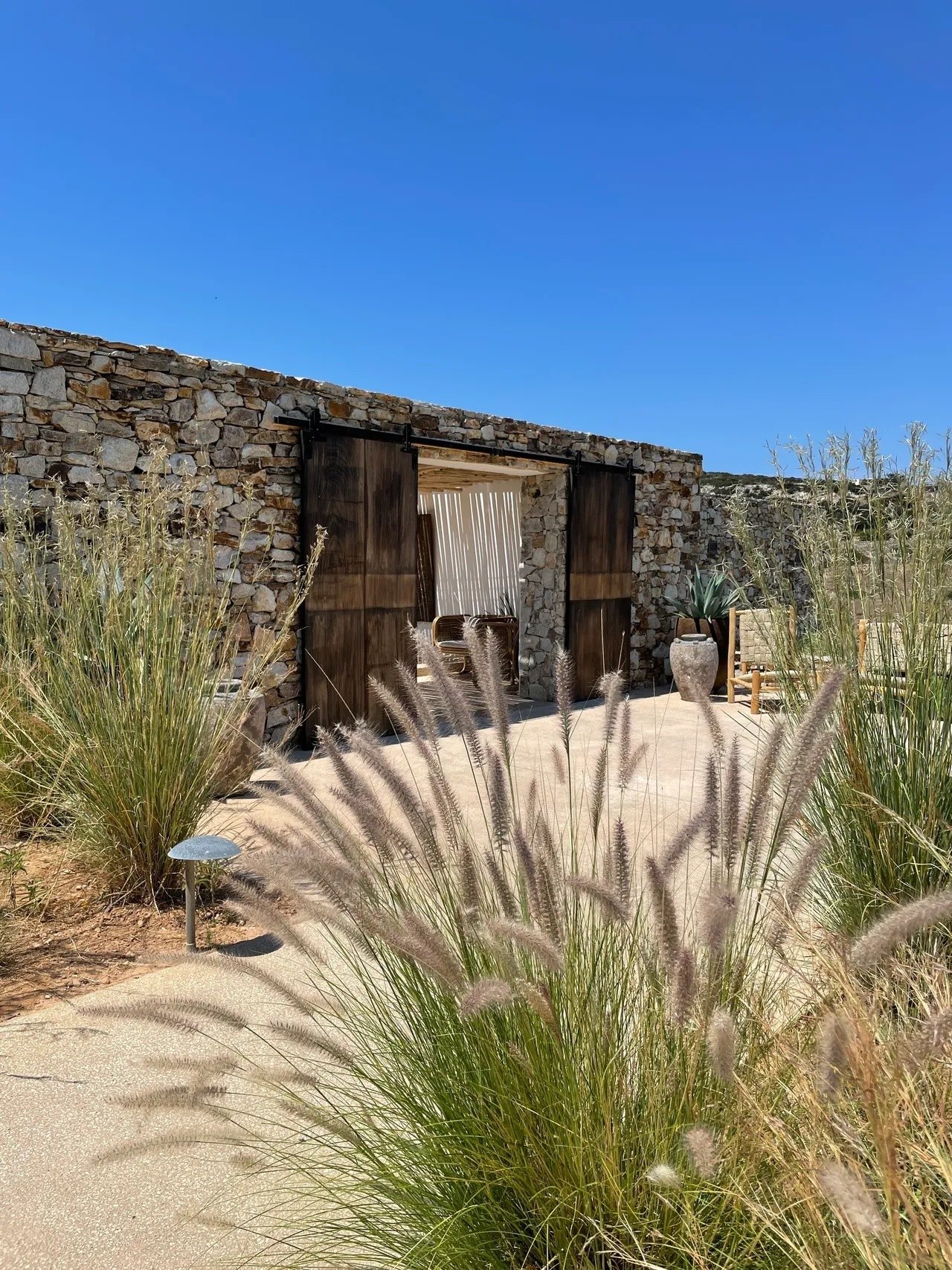 Tall timber doors in a dry-stone wall at The Rooster at blue hour, with feathered pennisetum grasses in front and a terracotta urn to one side