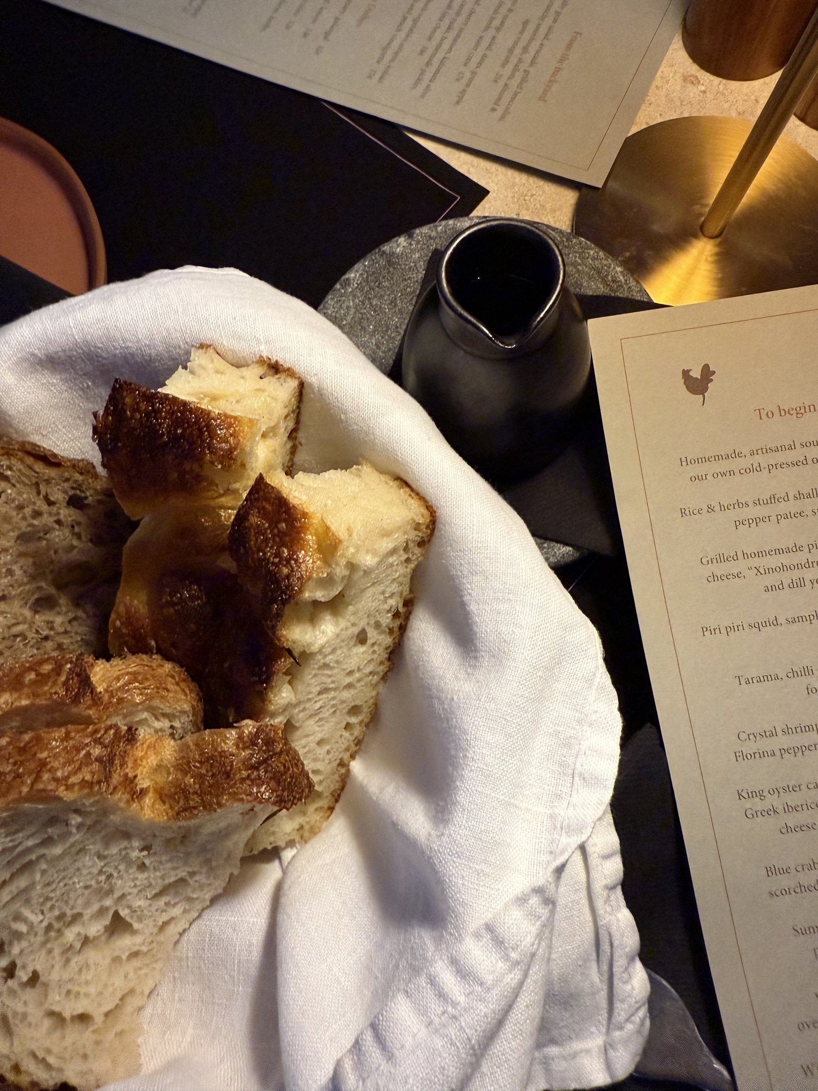 A white cloth opened on a table at The Rooster holding torn pieces of pale crusted bread, beside a black carafe and a printed menu