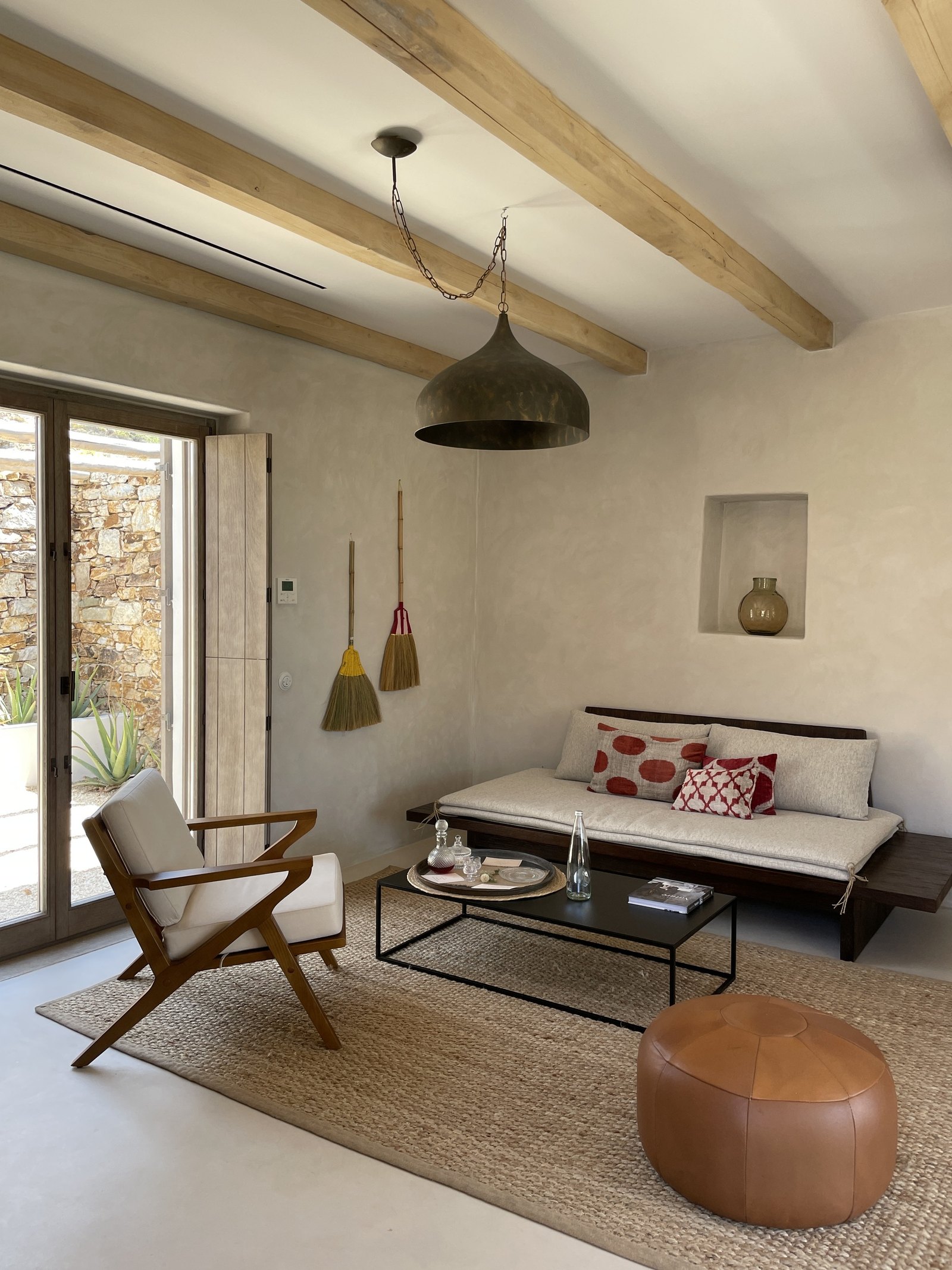 A sitting room at The Rooster with a low daybed, a timber armchair, a leather pouf and a broom standing against the white wall by the door