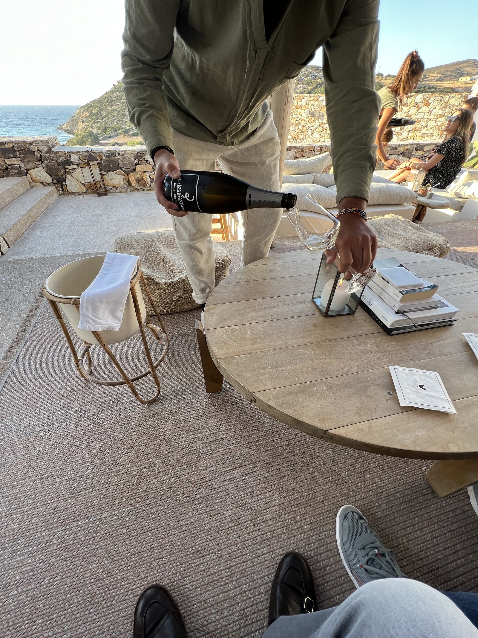 A member of staff in a green shirt leaning over a round timber table at The Rooster to pour a bottle of Billecart-Salmon brut into a glass, with a cooler and a lantern on the table and the bay and headland behind