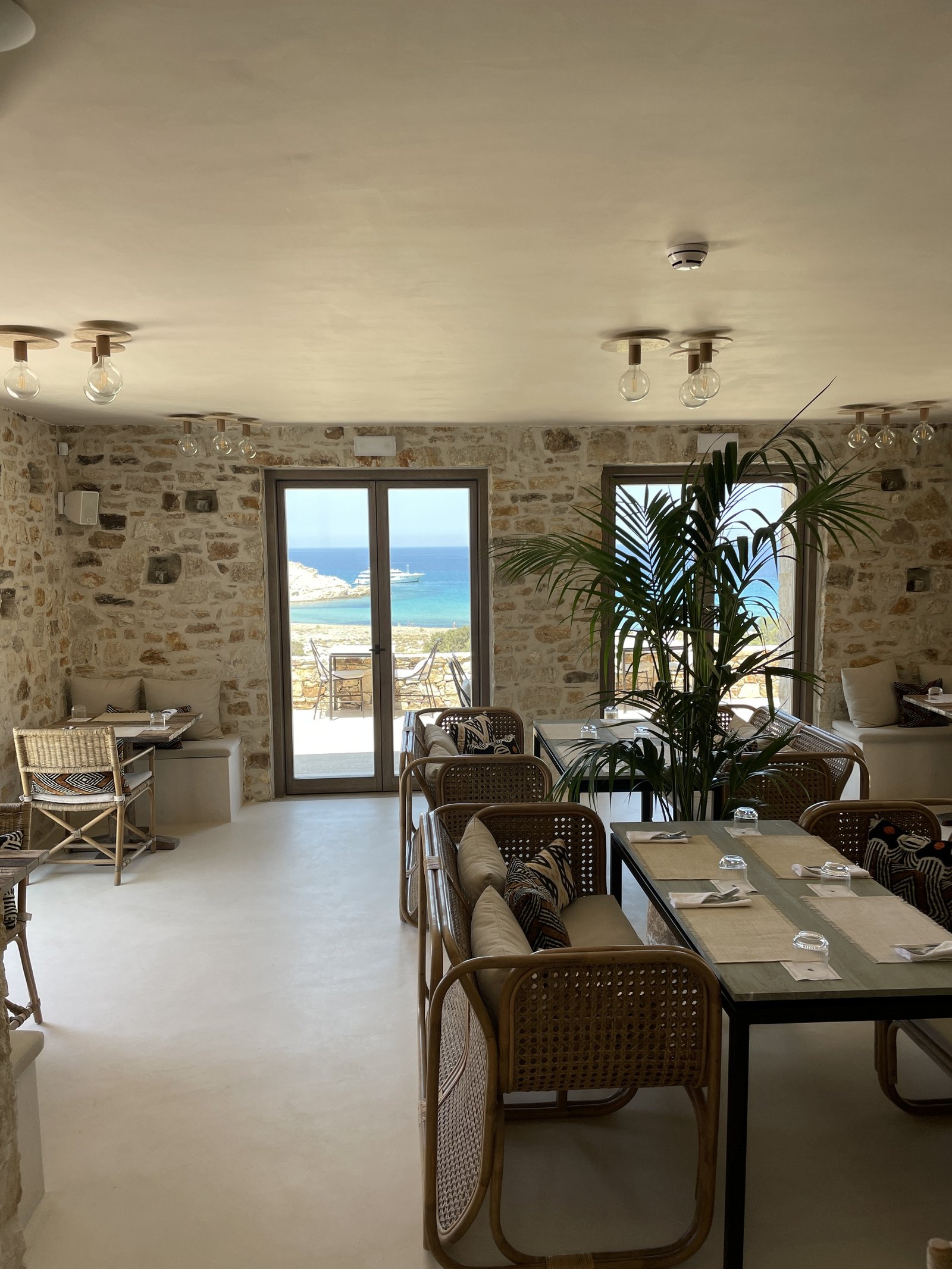 The dining room at The Rooster with stone walls, cane chairs and timber tables, open through tall doors to the bay and a palm