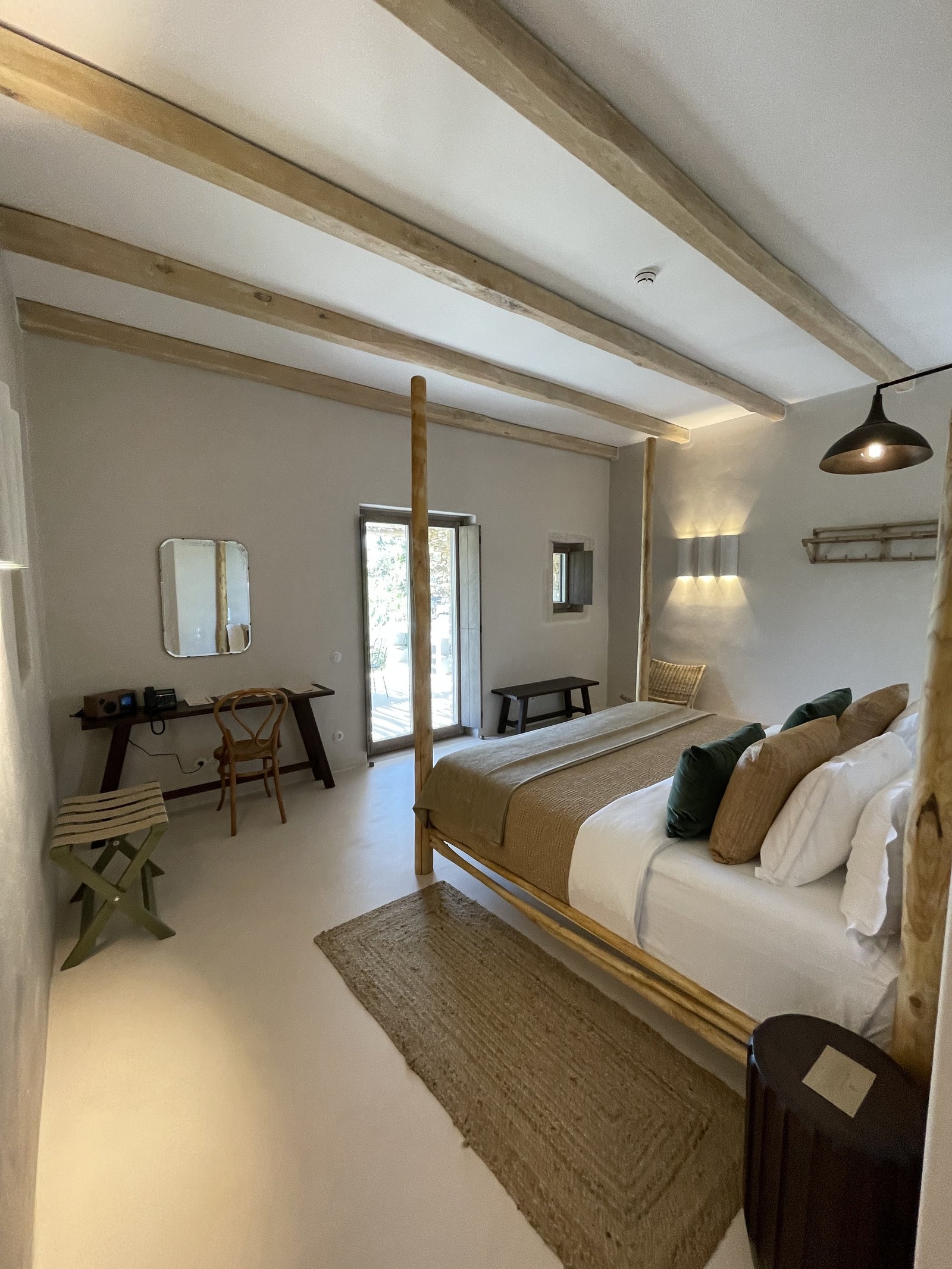 A bed with a four-poster frame of pale driftwood at The Rooster under a beamed ceiling, with a small mirror and a folding stand against the wall