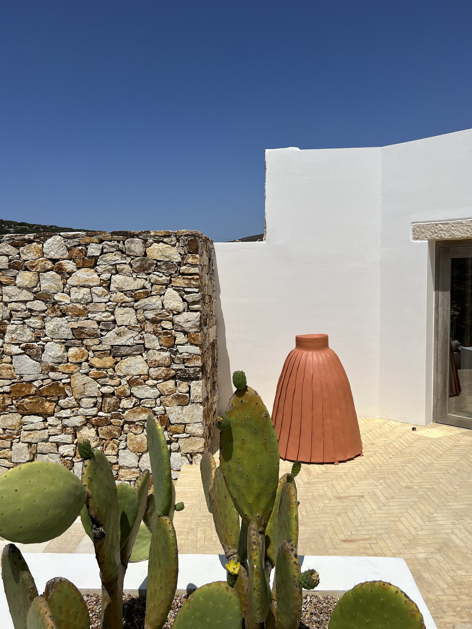 A large terracotta lamp standing in a bed of prickly pear against a white wall and a dry-stone wall at The Rooster, under a clear sky
