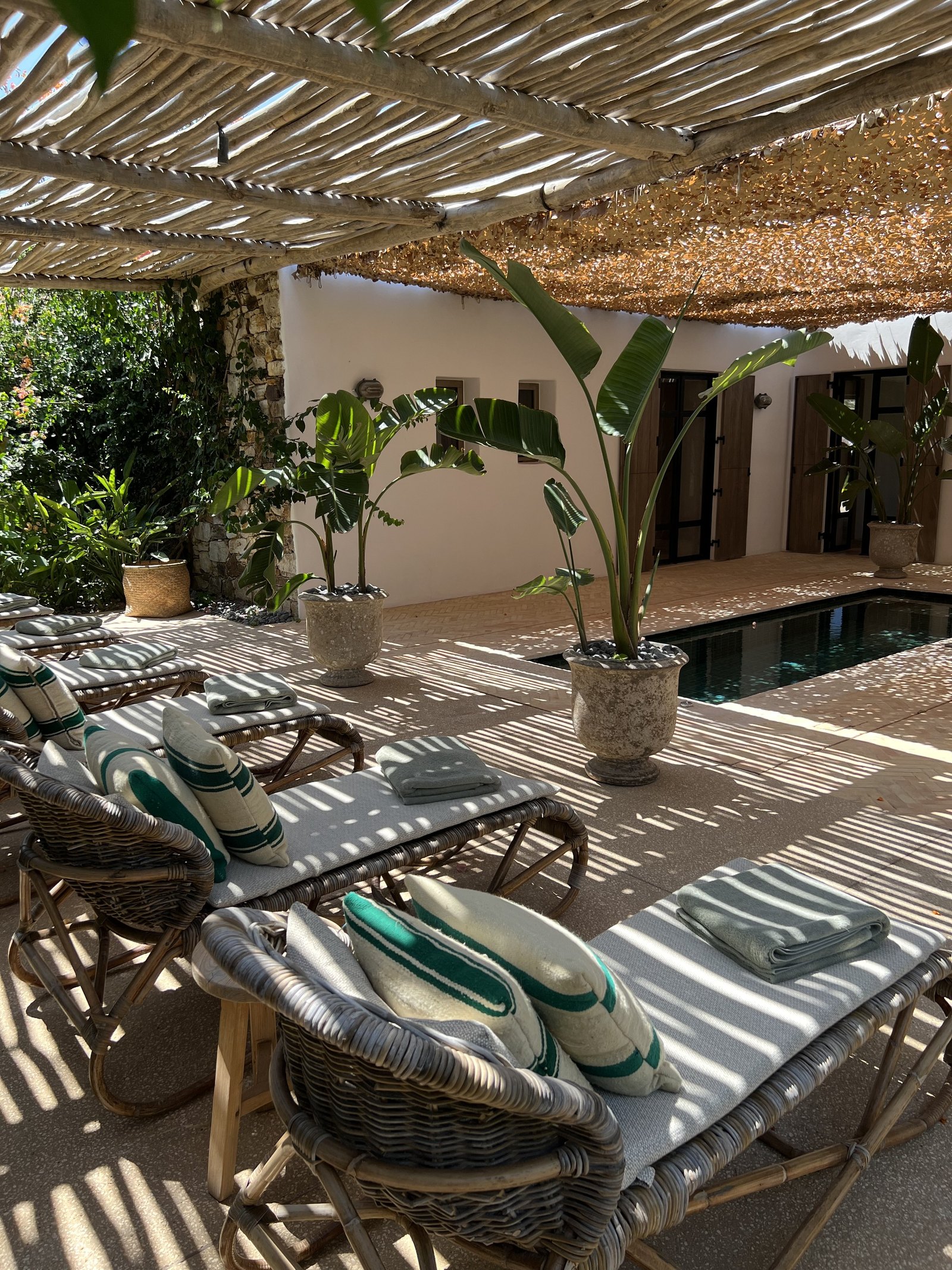 Cane loungers at The Rooster with striped towels folded on them and slippers beside, under the dappled shade of the woven awning