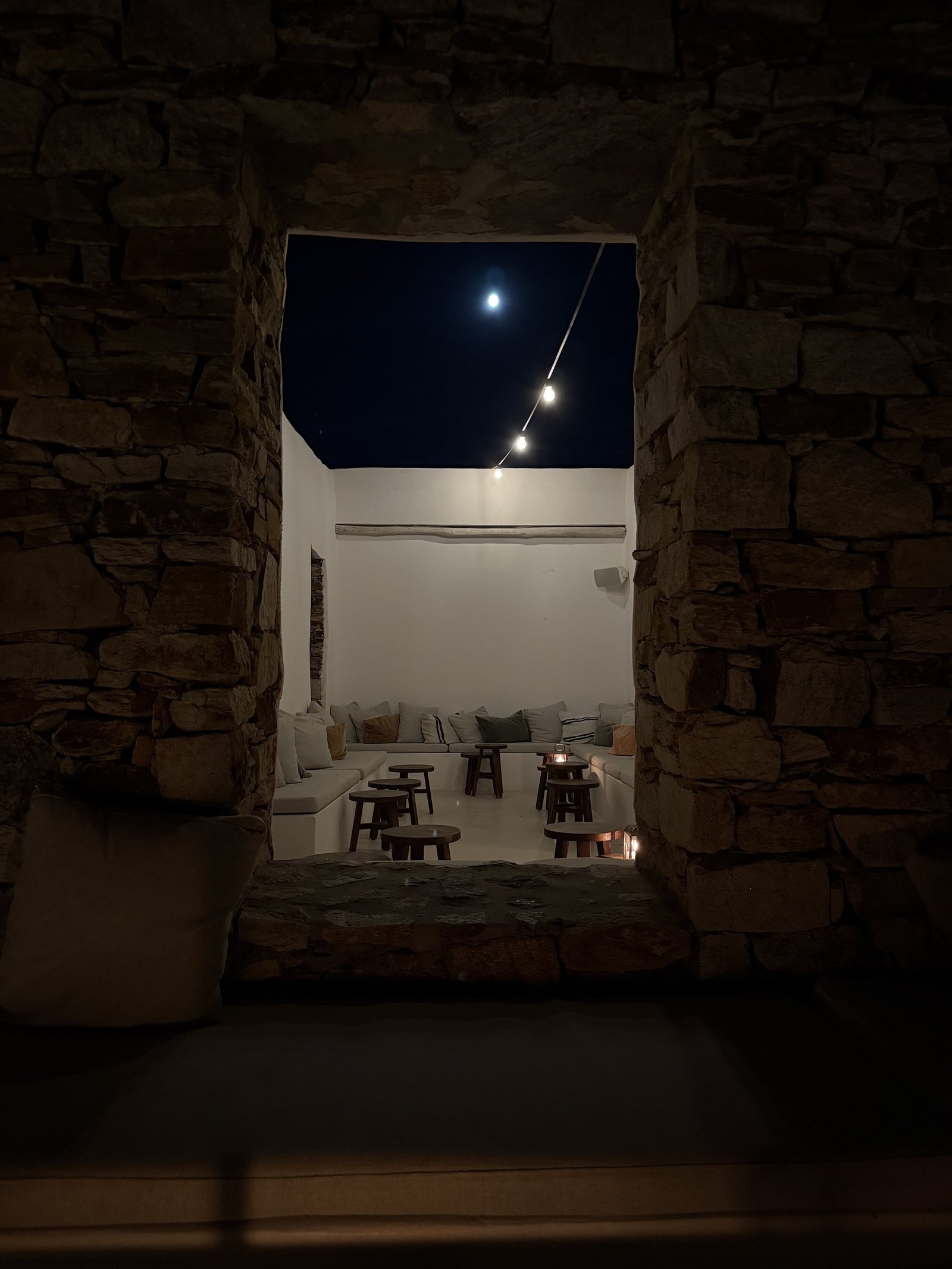 The sunken lounge at The Rooster at night, lit low between rough stone walls, with the moon in the strip of dark sky above it
