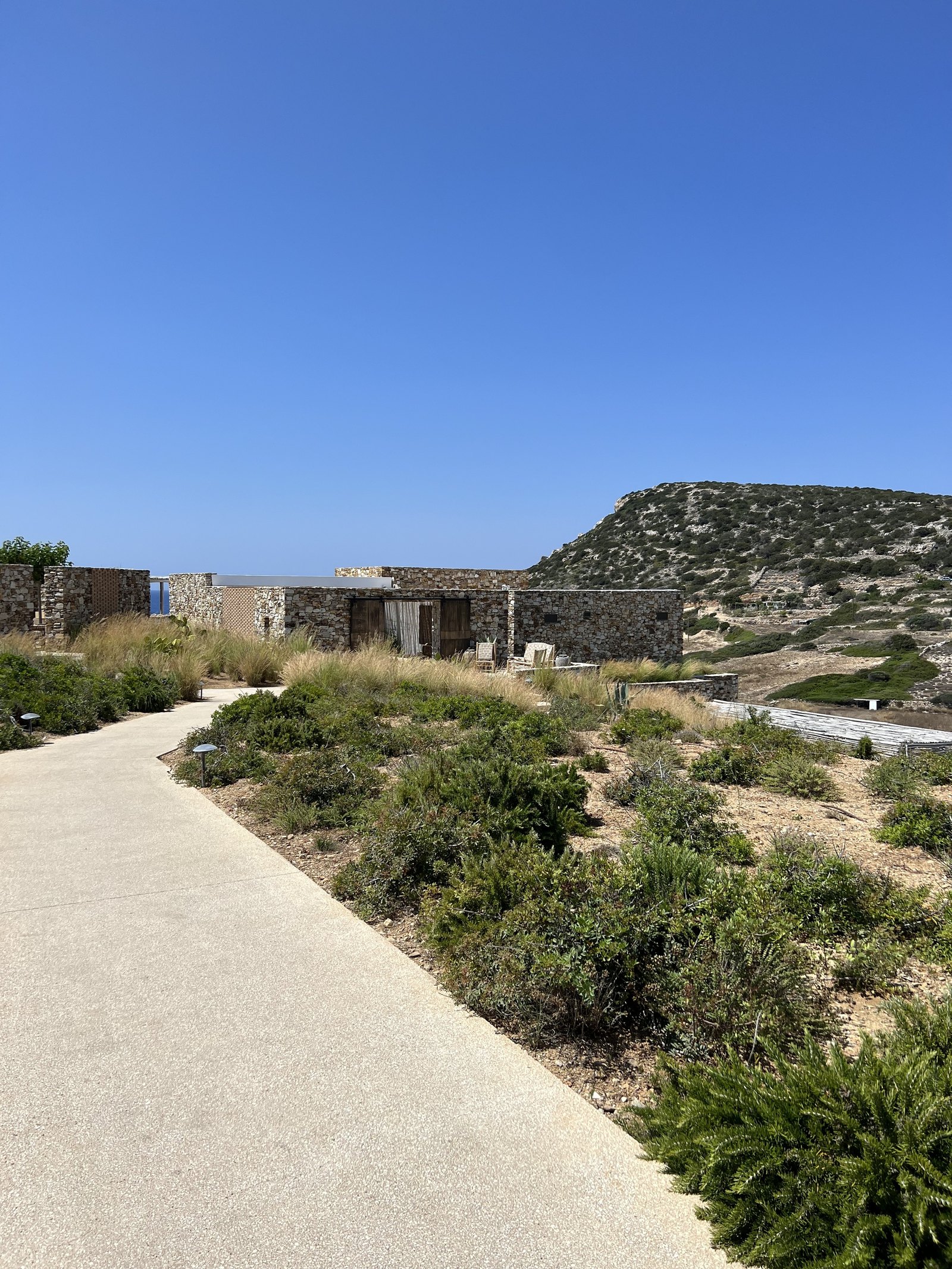 A pale gravel path at The Rooster running through low scrub towards long stone buildings, with a flat headland and the sea behind