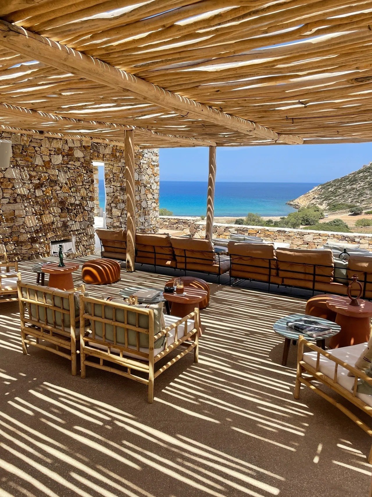 A bamboo pergola over low cane and leather seating at The Rooster, open to the turquoise water of Livadia Bay and the headland beyond