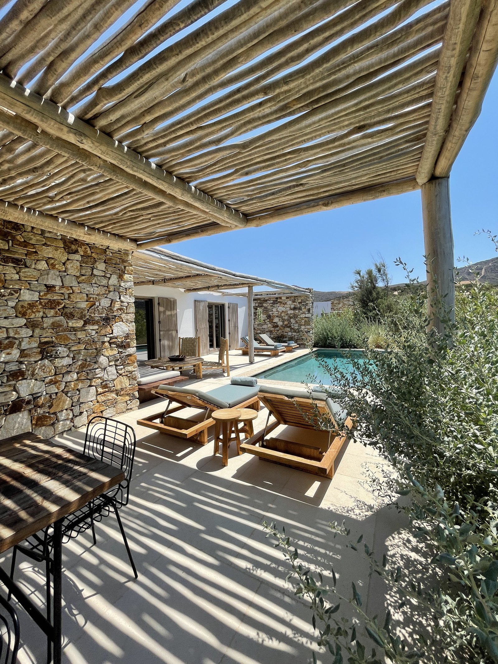 A terrace at The Rooster under a pergola of rough timber poles throwing hard striped shadows across the stone and the loungers