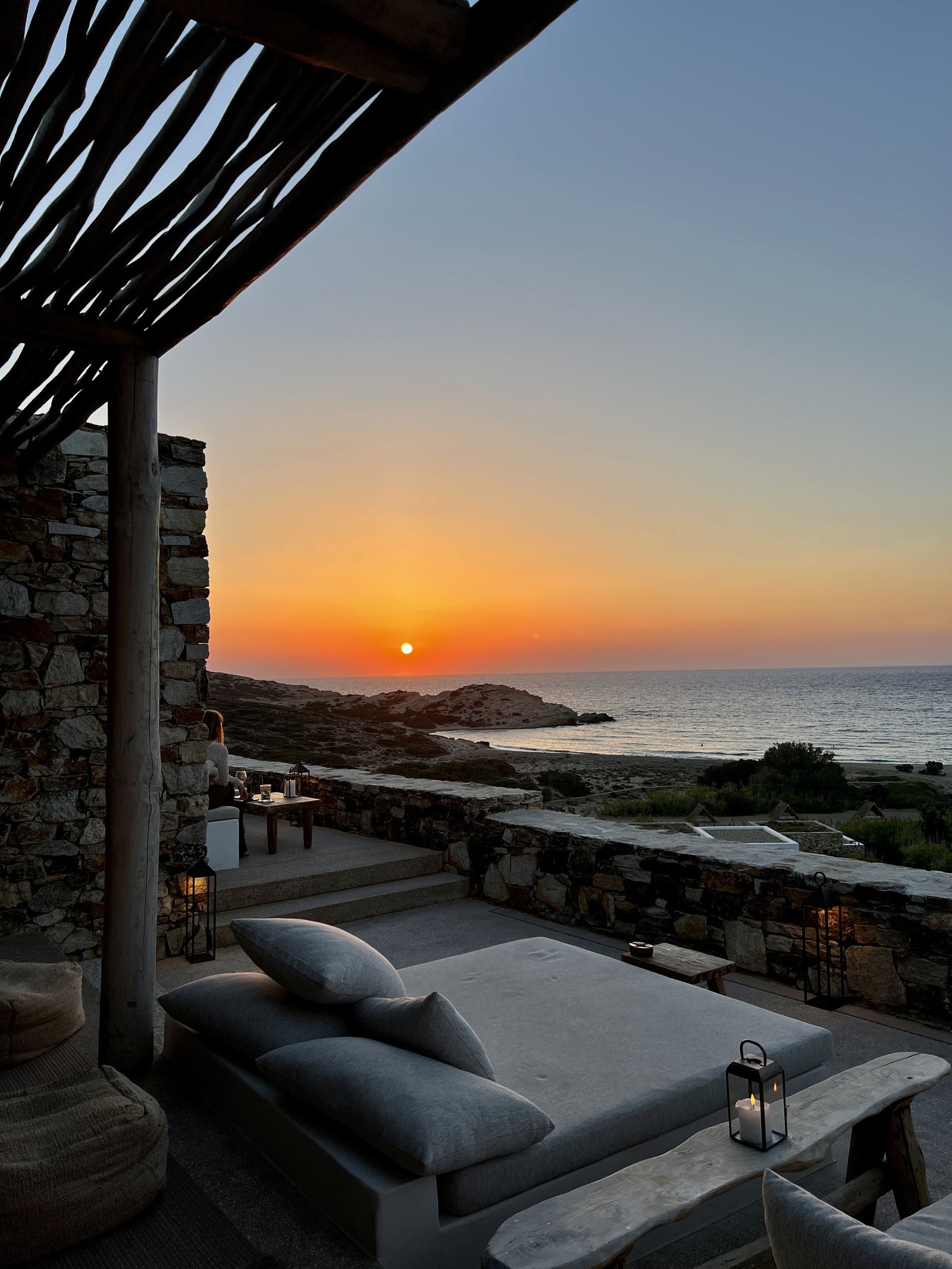The sun dropping into the sea beyond Livadia Bay seen from a stone terrace at The Rooster, with cushions on a low wall and lanterns in the foreground