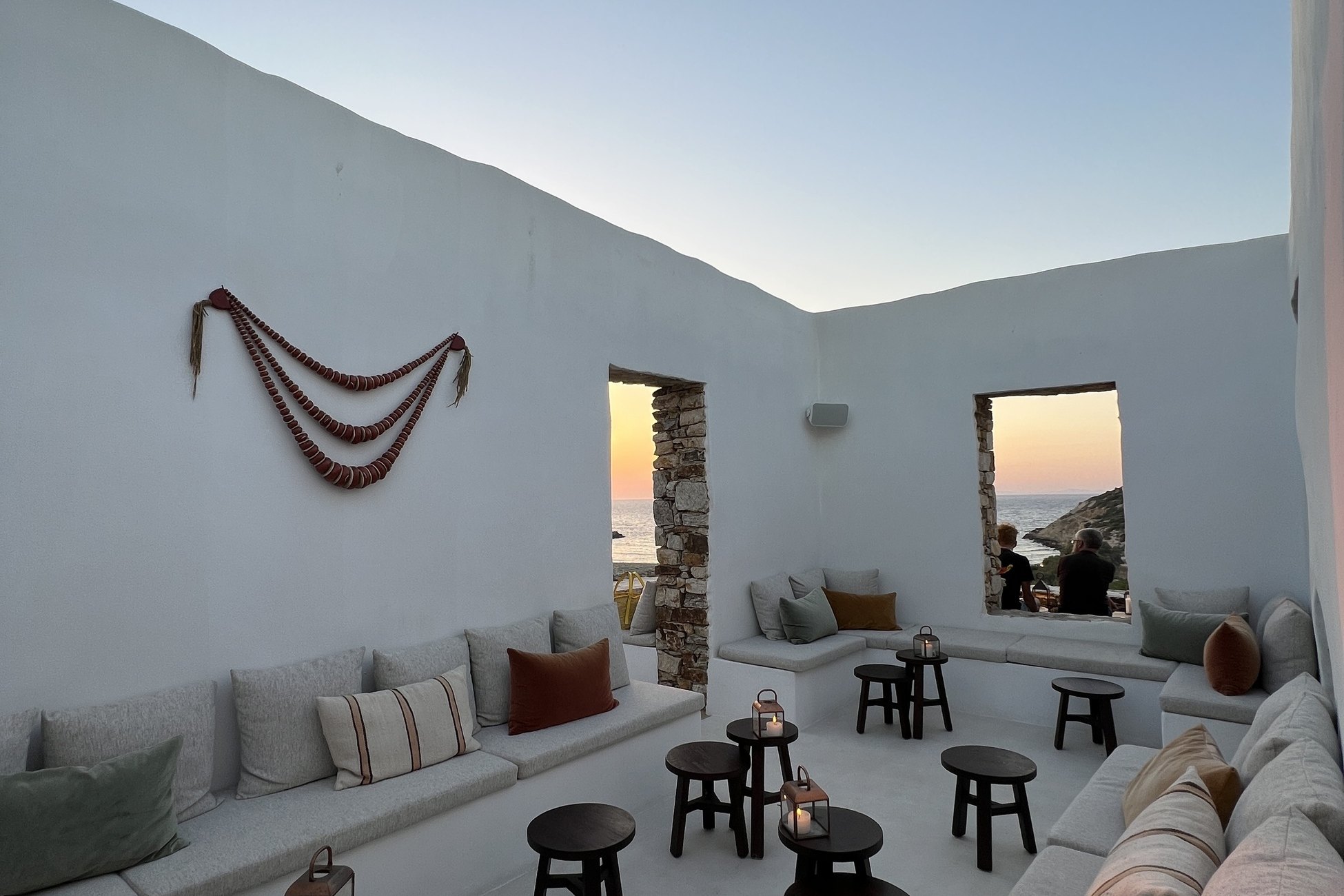 A sunken white courtyard at The Rooster lined with long built-in benches and cushions, with small black stools set in front of them and openings in the wall onto the sea