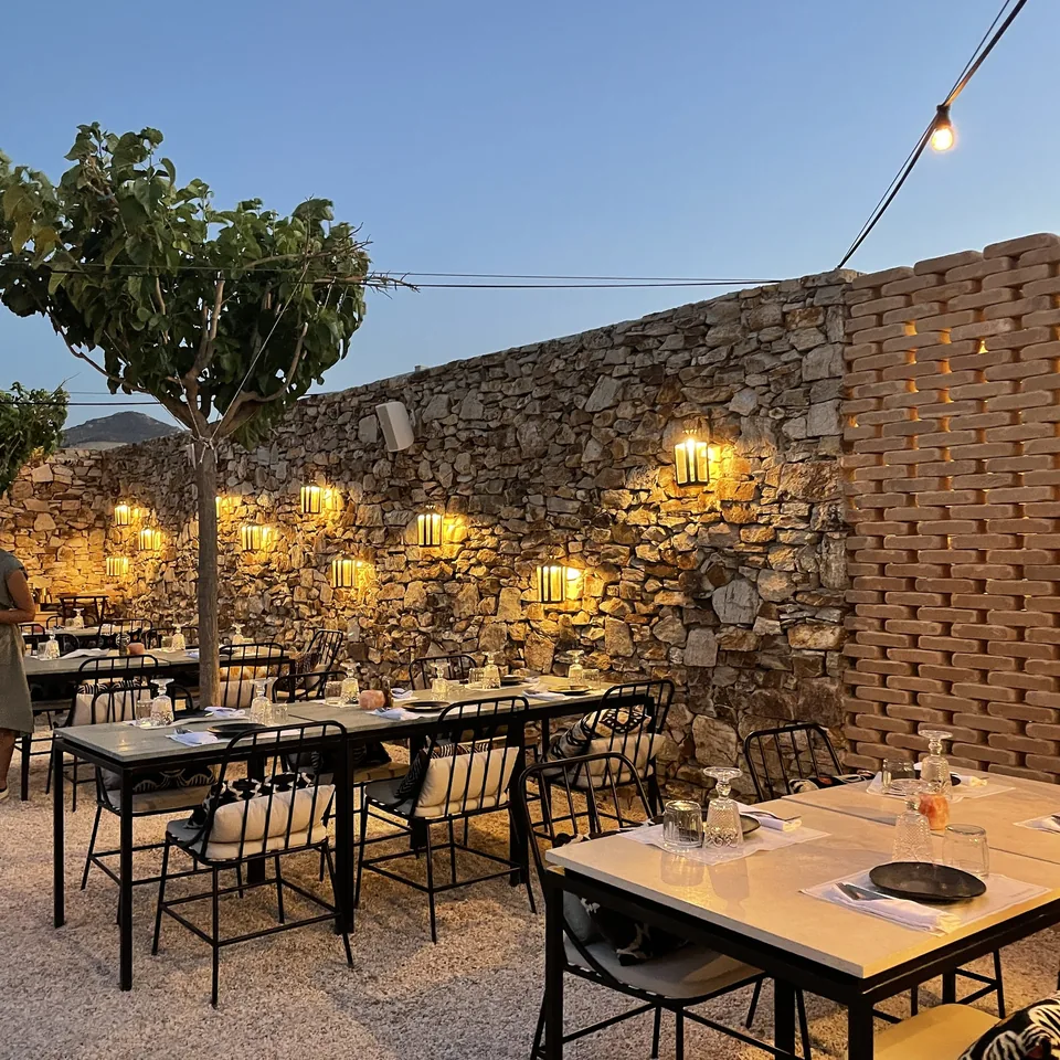 Tables laid on gravel against a lit dry-stone wall at The Rooster at dusk, with a small tree and lanterns along the wall