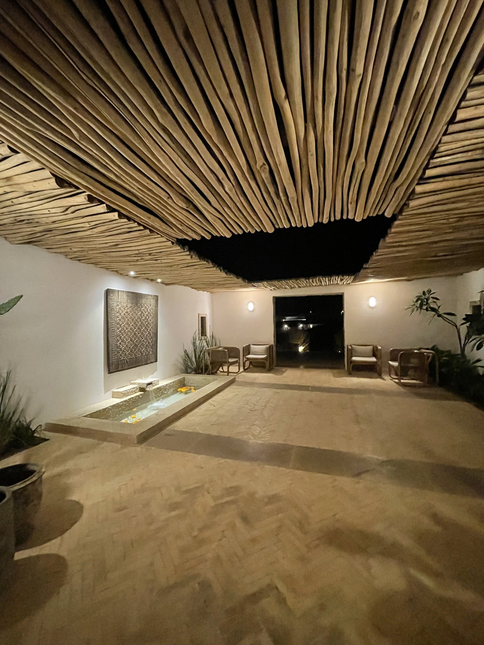 A room at The Rooster under a dark thatched ceiling with a shallow sunken basin of water in the floor and plants in the corners
