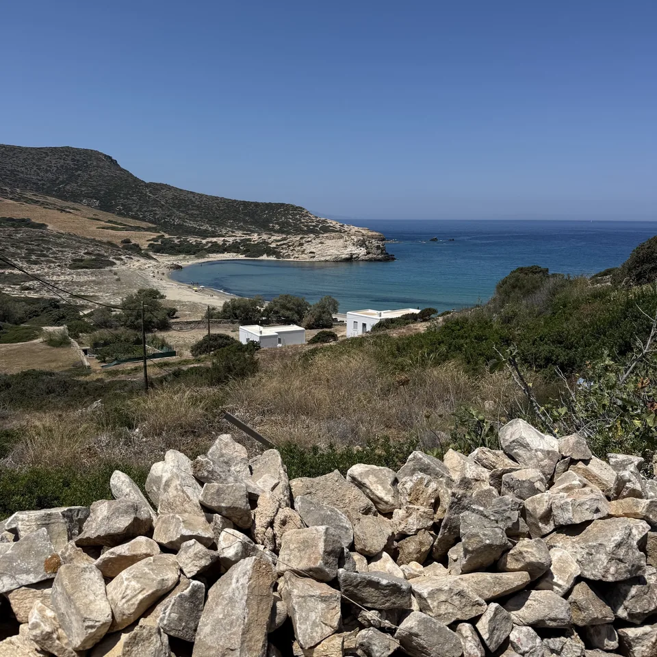 Livadia Bay seen from the road above, with a pile of dry stones in the foreground, the low white buildings along the shore and the headland behind