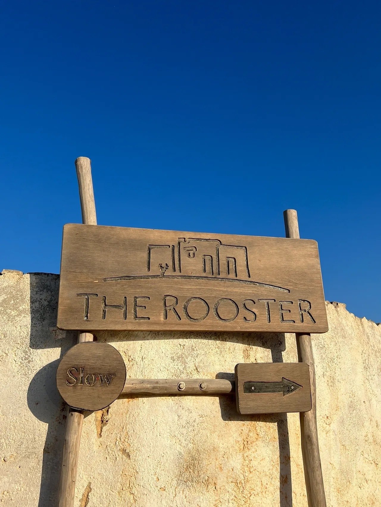 A weathered timber sign at The Rooster carved with the hotel's name and the word Slow under an arrow, against a deep blue sky