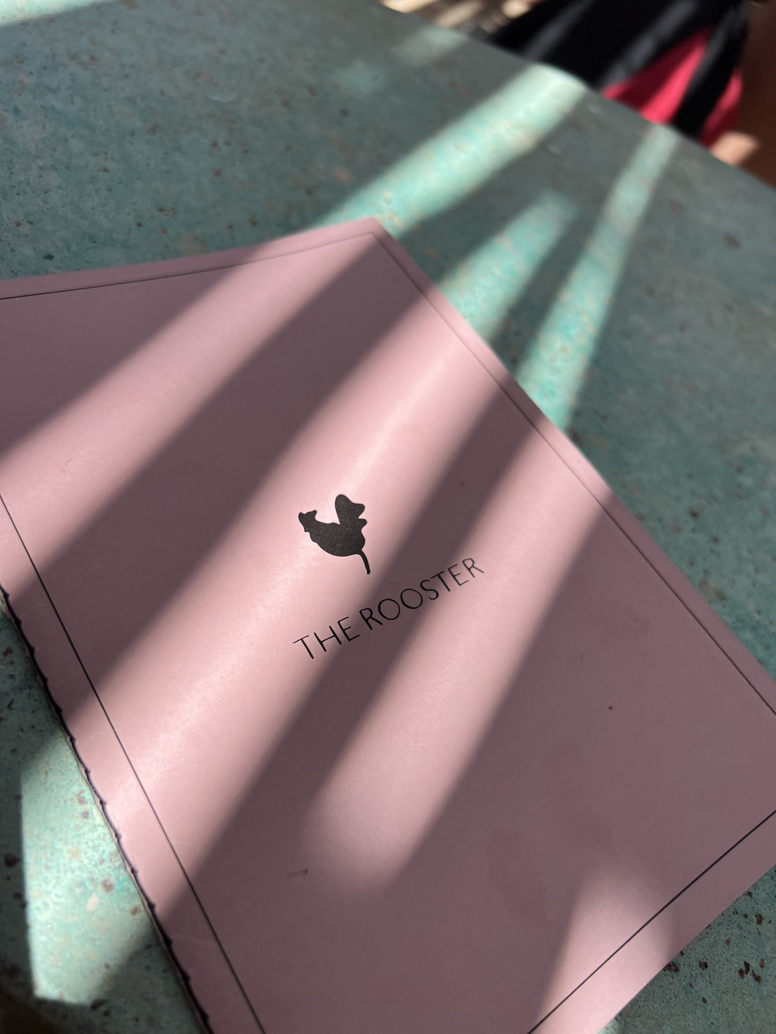 A soft pink menu card at The Rooster printed with a small rooster mark and the hotel's name, lying on a stone table in dappled shade