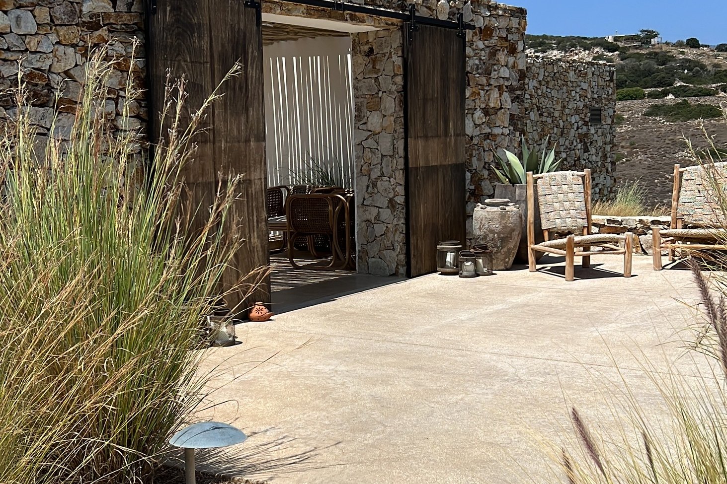 The entrance at The Rooster, a gap in a dry-stone wall closed by tall timber doors, with tall feathered grasses in front of it and the sea beyond