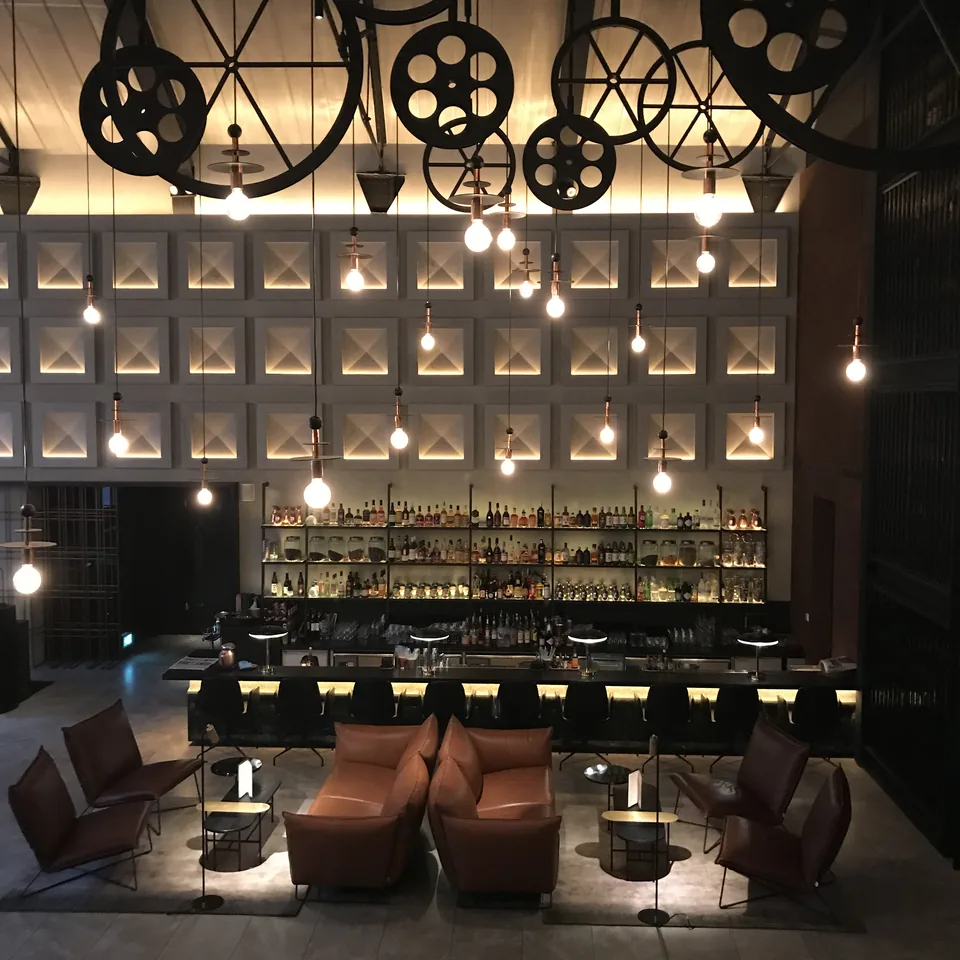 A long lit bar counter at night under hanging pulley wheels, with a wall of bottles behind it