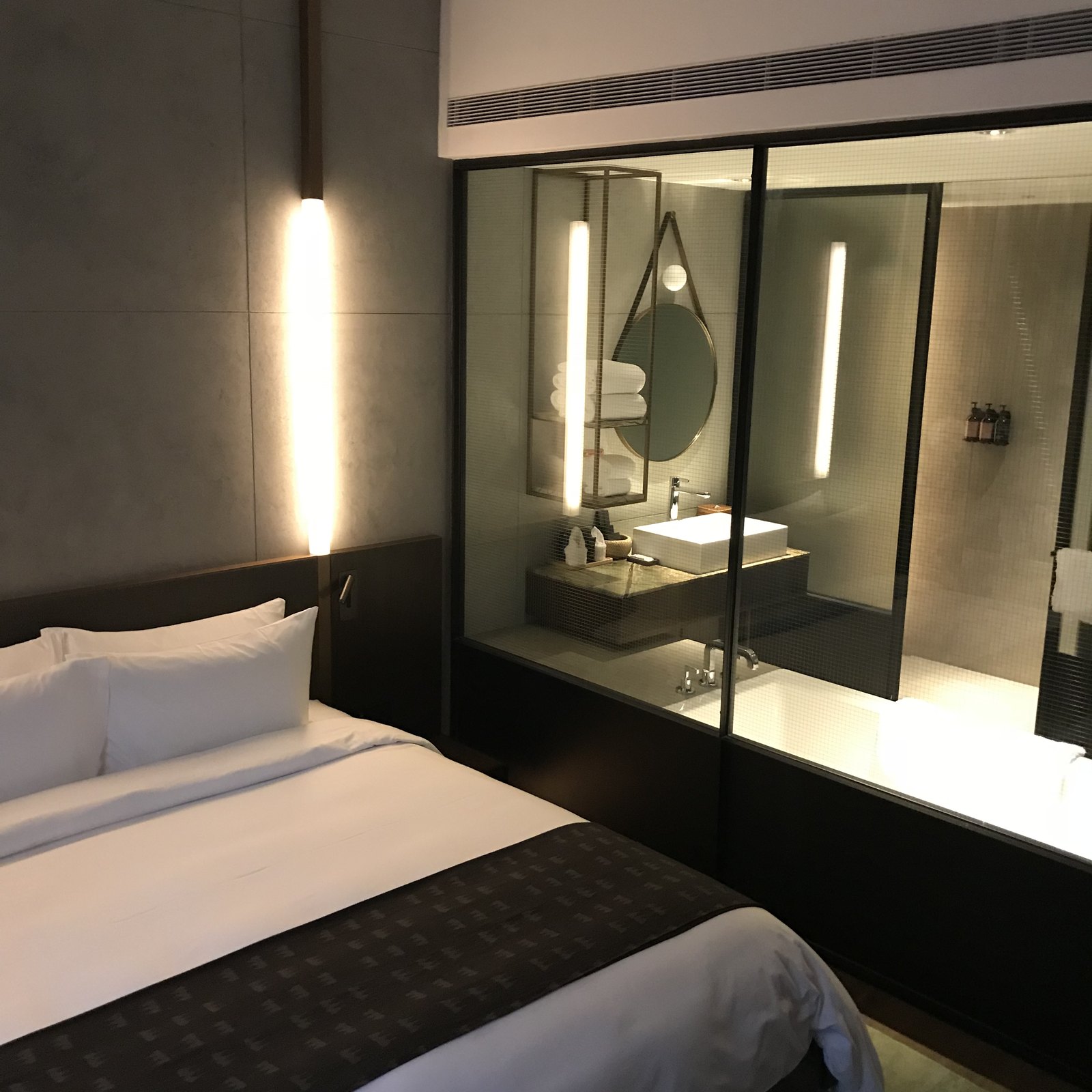 A made bed with a dark runner, a vertical light strip on the wall and the lit bathroom through a glass partition