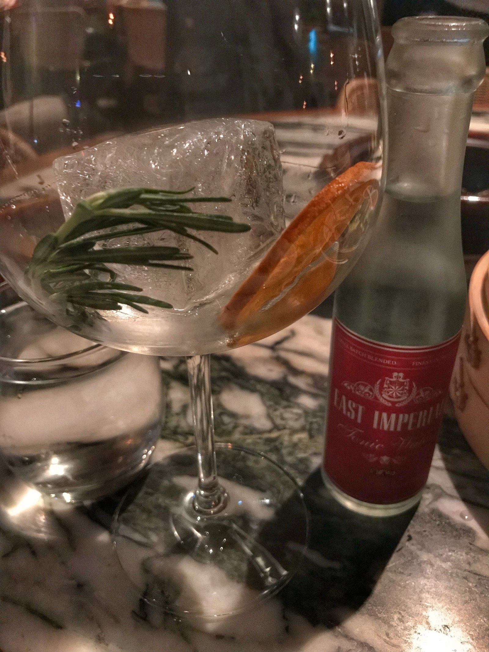 A coupe glass with a long drink and a garnish beside a small bottle with a red label on marble