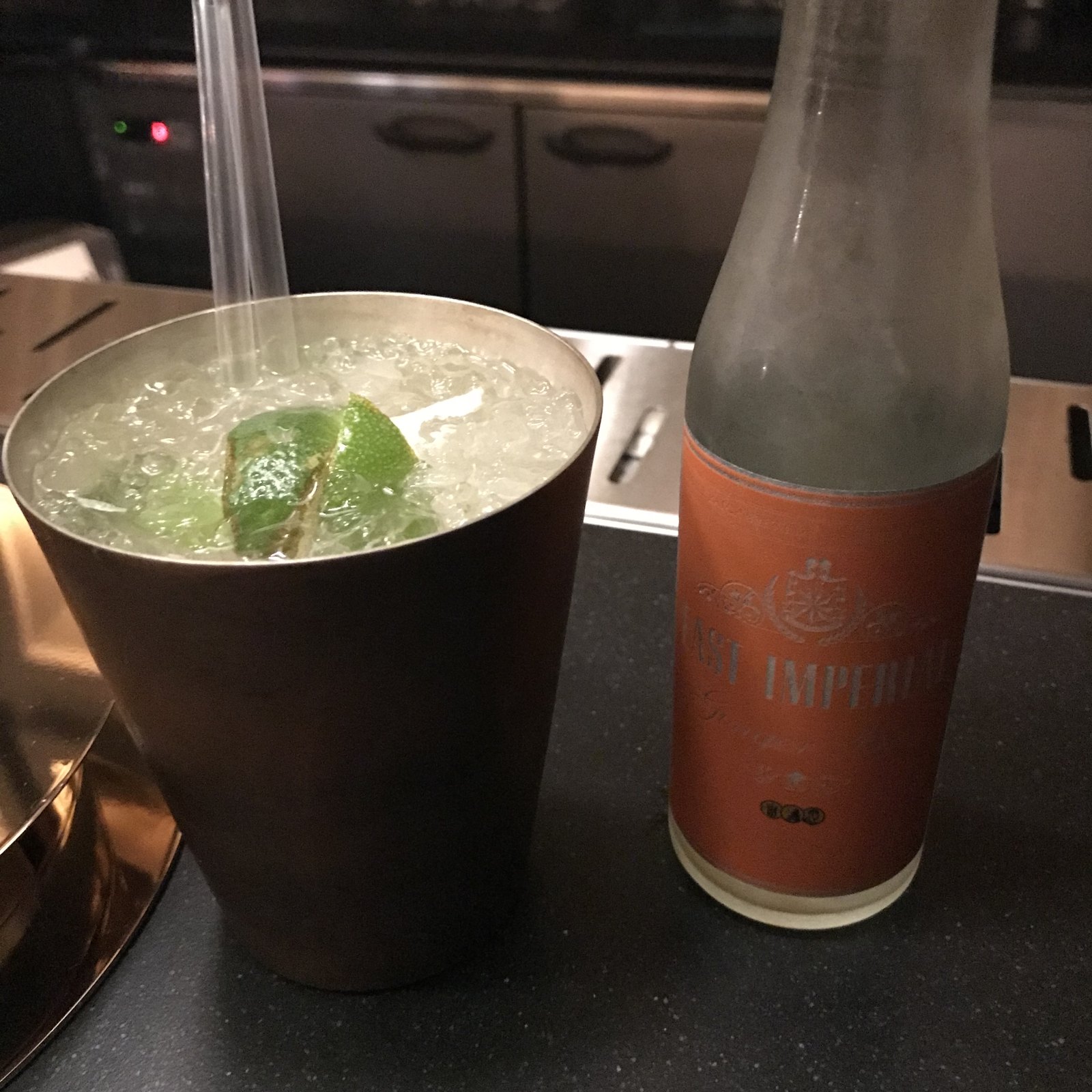 A tumbler with a lime wedge and a straw beside a tall bottle on a dark counter