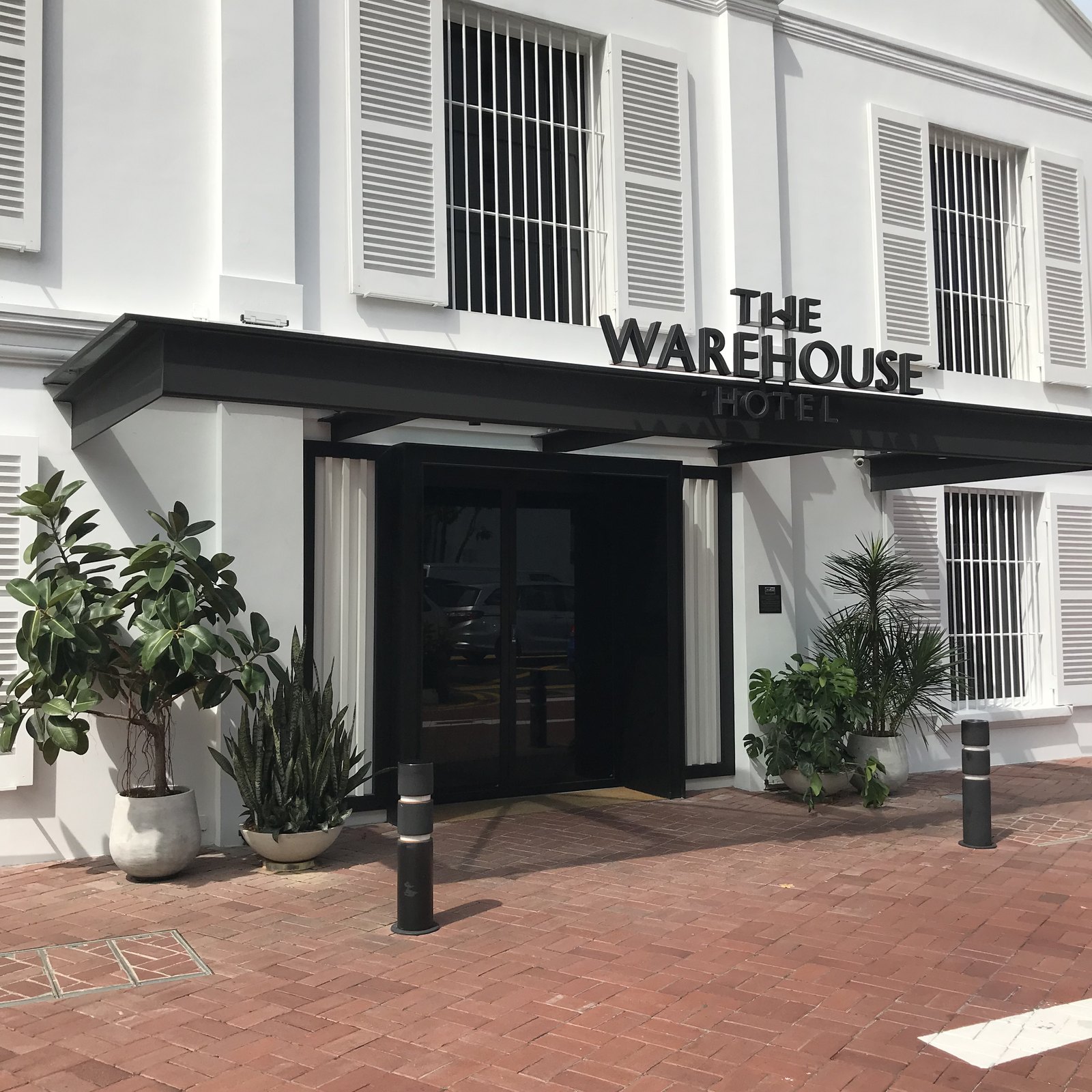 A white godown front on Havelock Road with THE WAREHOUSE HOTEL lettered across a black steel canopy over the doors, planted pots on brick paving