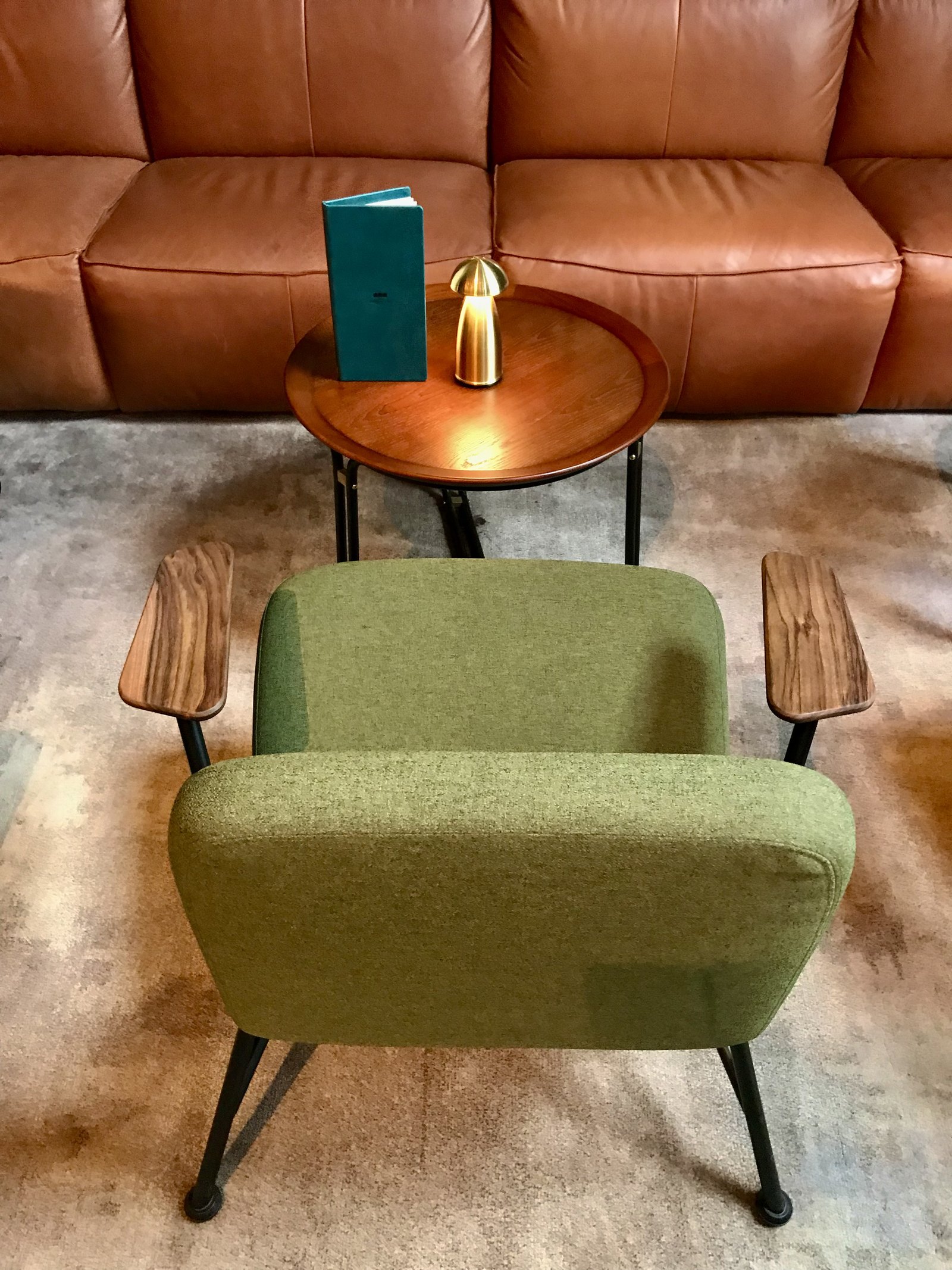 A green upholstered armchair with timber arms in front of a tan leather sofa and a brass lamp