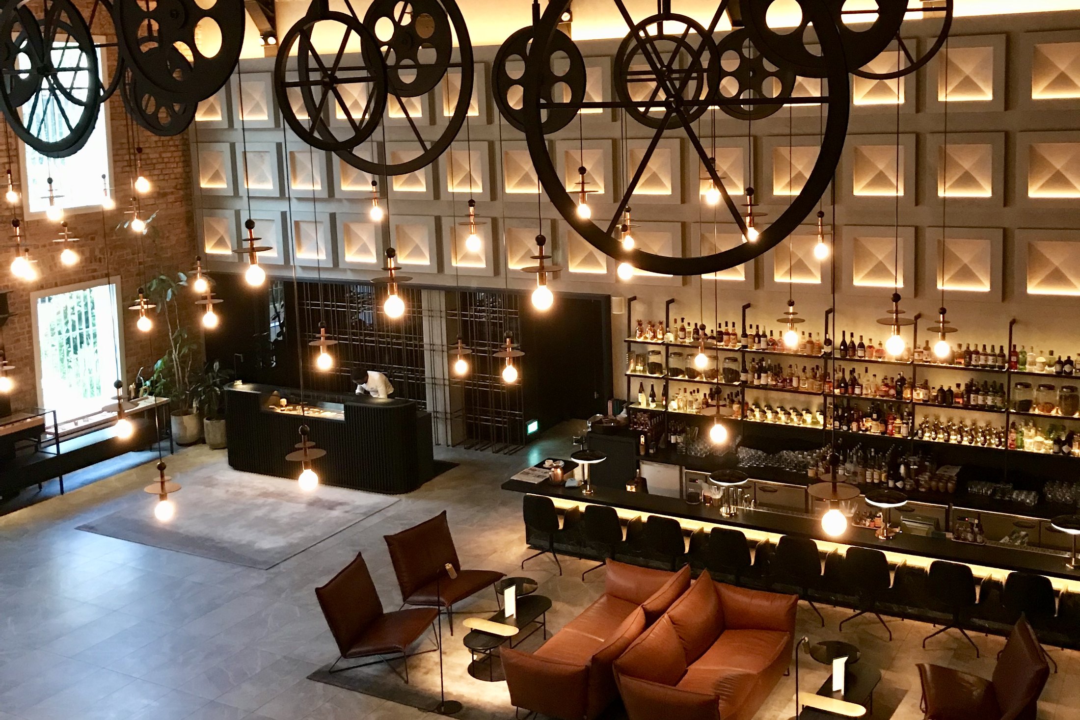 A tall lobby bar at night, cast-iron pulley wheels hanging on rods under the roof trusses, a lit wall of bottles down one side and leather chairs below