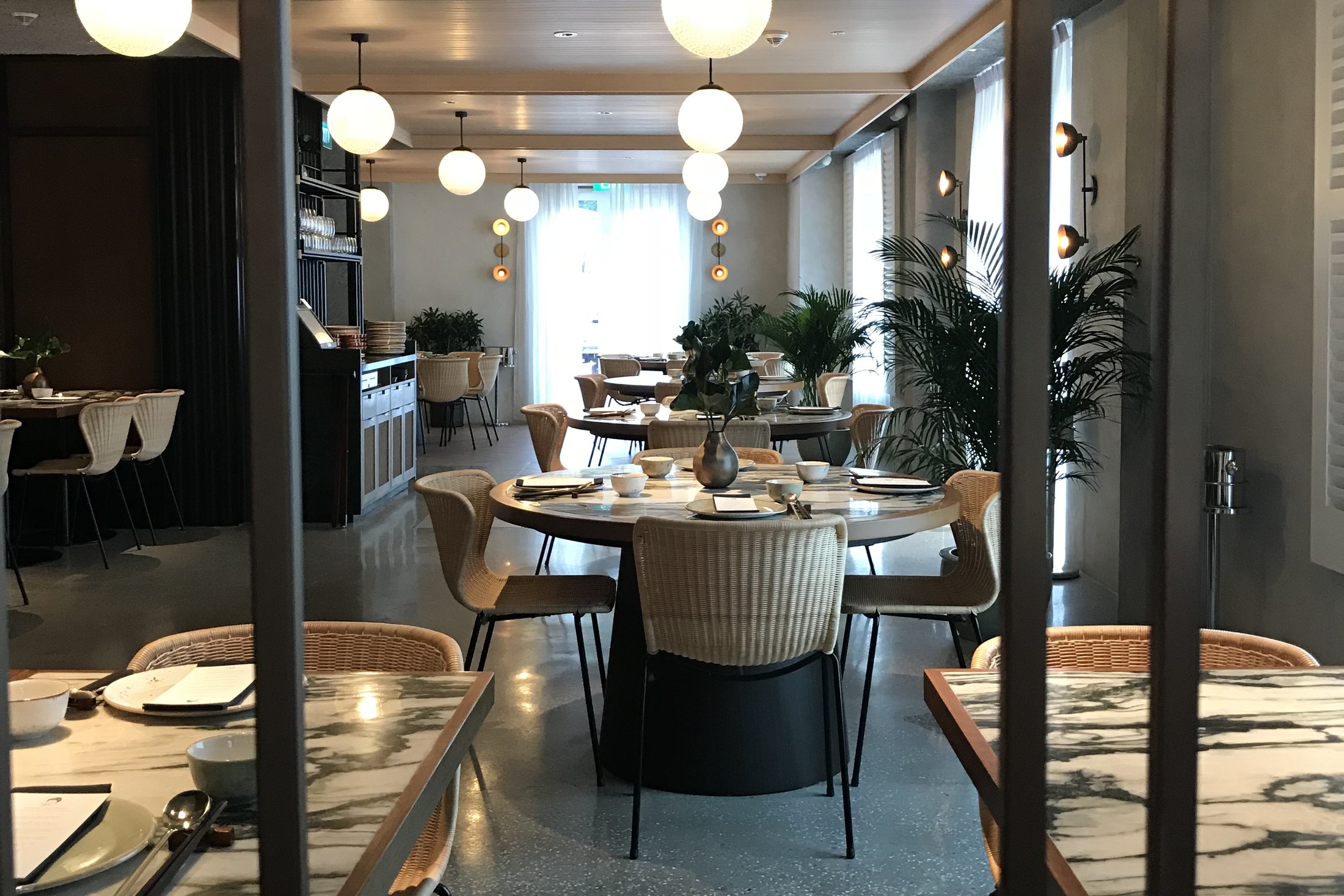 A dining room seen through a black steel frame, round tables laid with marble tops, rattan chairs and white globe lights down the length of the room