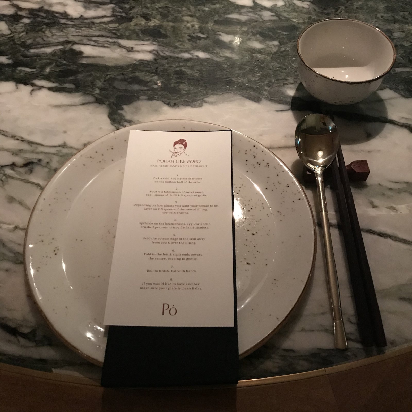 A place setting on marble with a speckled plate, chopsticks and a printed menu card reading Po