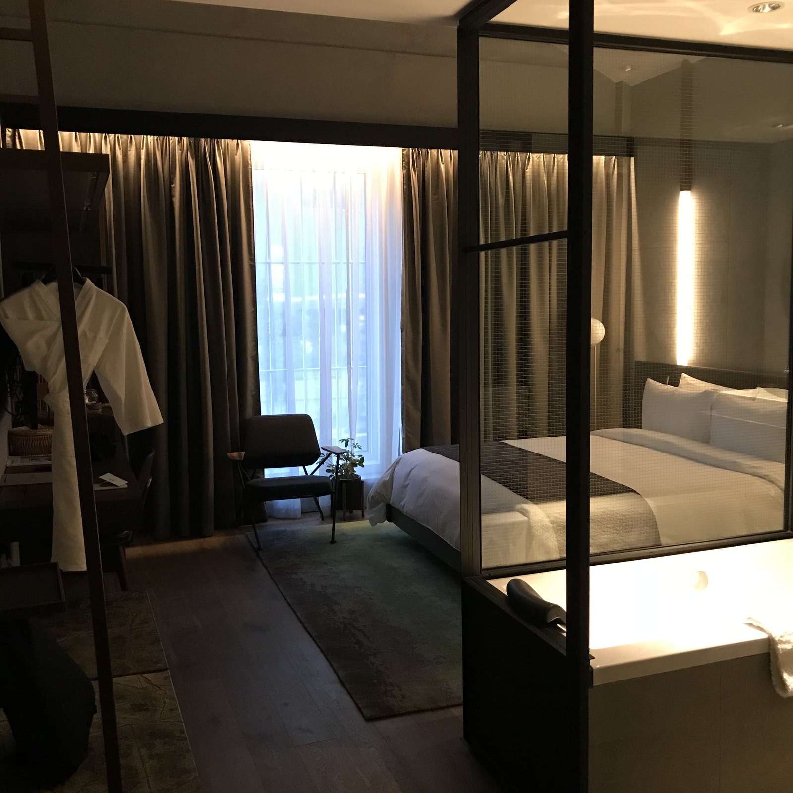 A bedroom with a black steel four-poster frame around the bed, a glazed partition to the bathroom and a curtained window beyond
